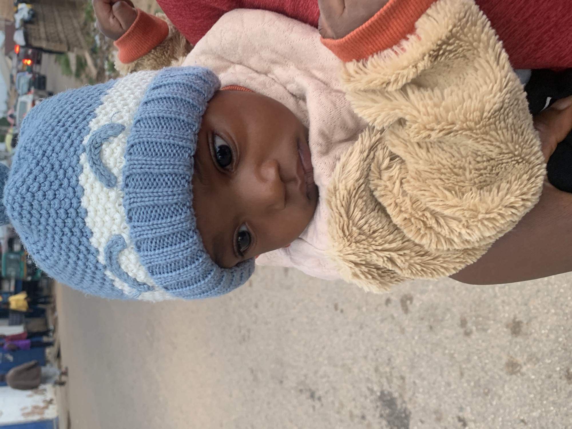 A baby is being held in someone's arms in an outdoor location, possibly in Jos, Nigeria. The baby is wearing a blue and white knit hat and a tan fuzzy jacket. The person holding the baby is wearing a red shirt with orange cuffs. The background appears to be a street scene with buildings and possibly traffic. The ground is paved.
