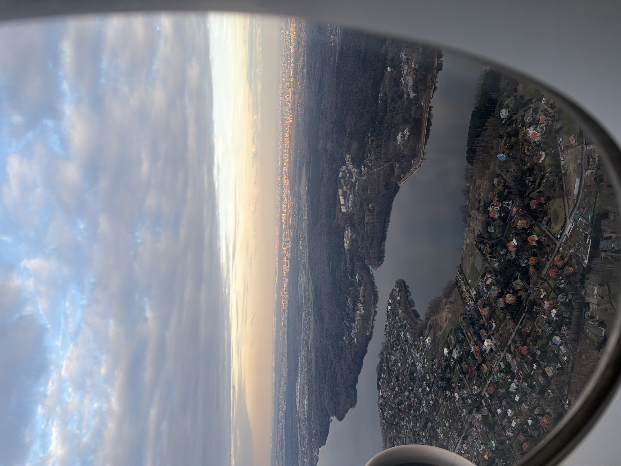 The image shows a view from an airplane window. On the left, there is a cloudy sky with the sun setting on the horizon. In the center, a city skyline is visible, merging with the edge of a large forest. A river runs along the right side of the forest. On the right side of the frame, a residential area with many houses and streets is visible from above. The airplane window frame surrounds the view. Part of the plane's engine is visible in the lower right corner.