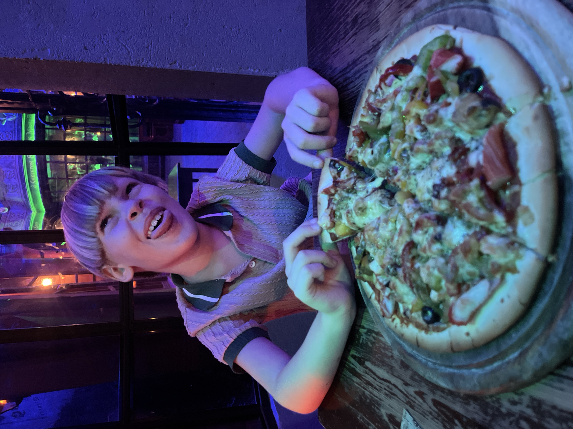 A young boy with a bowl cut is sitting at a wooden table, sticking his tongue out with a playful expression. In front of him is a whole pizza on a metal platter, with one slice already removed. He is wearing a collared shirt with a textured knit pattern. The background is dark with neon lighting, suggesting an indoor environment, possibly a themed restaurant or bar. The lighting in the scene is dramatic, with blue and pink hues illuminating the boy and the table. There is no discernible connection to Phuket City Municipality, Thailand, as the image lacks any recognizable landmarks or contextual clues to support such a location.
