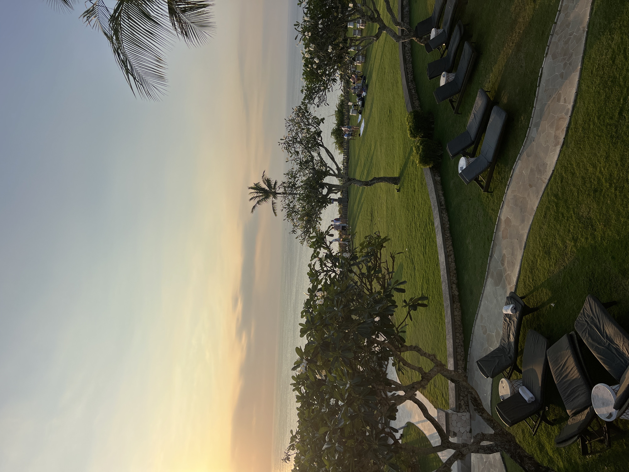 This image captures a serene sunset scene at a resort in Kuta Selatan, Indonesia. The foreground features several empty lounge chairs arranged on a well-manicured green lawn, some with white towels neatly placed on them. A winding stone pathway is visible, leading through the grass. In the mid-ground, there are more lounge chairs and groups of people gathered on the lawn, some appearing to be picnicking. Trees with white flowers are scattered across the landscape, and a lone palm tree stands tall against the horizon. The background is dominated by the calm ocean reflecting the warm hues of the setting sun. The sky transitions from a soft yellow and orange near the horizon to a pale blue. The overall atmosphere is peaceful and idyllic, indicative of a tropical evening.
