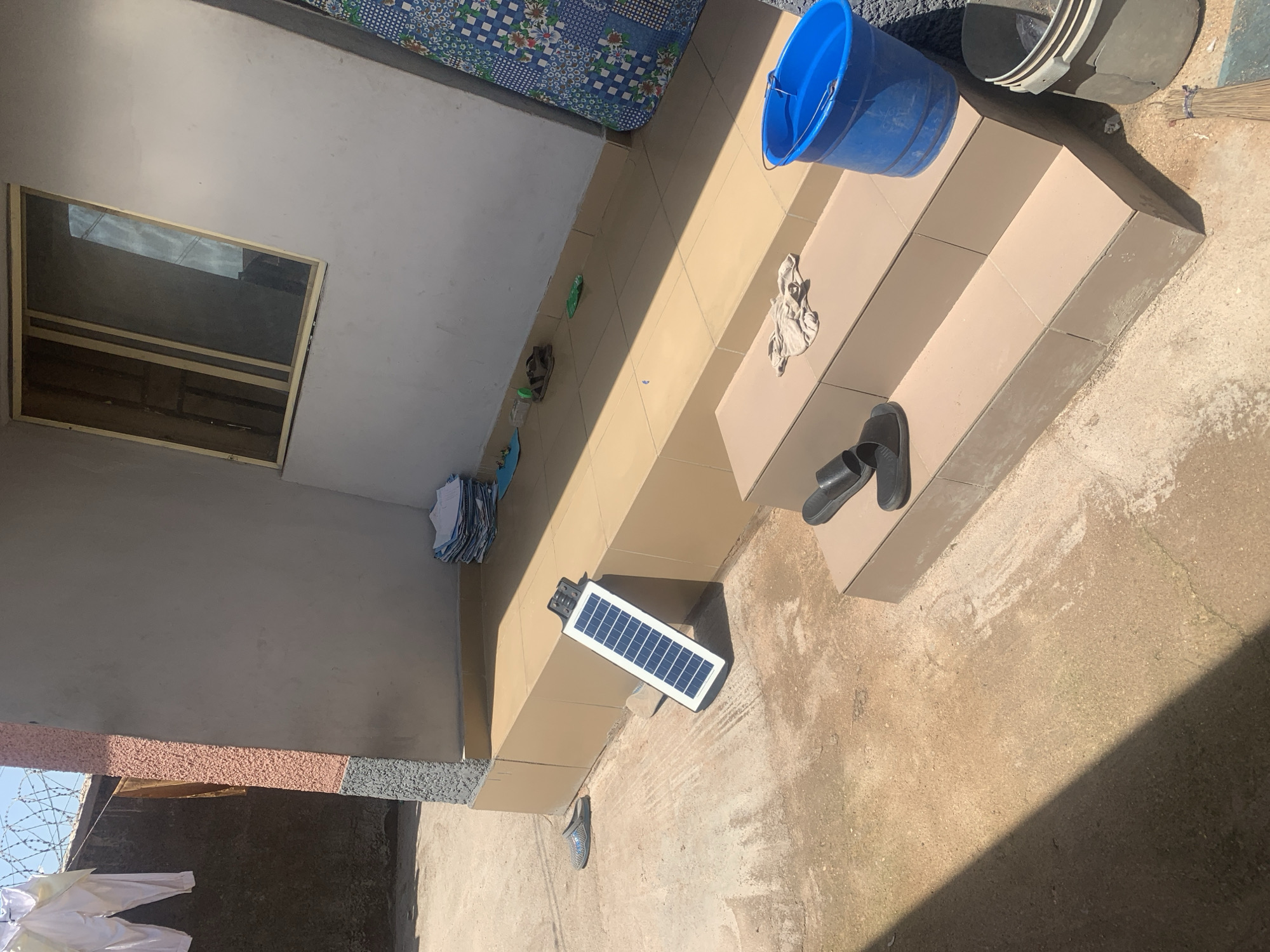 In Jauro Yinu, Nigeria, a solar panel rests on a tiled step outside a building. Two black flip-flops are positioned on a step below the solar panel. A blue bucket and a gray bucket containing what appears to be water and debris are located on the right side of the steps. A pile of clothes hangs from a wire on the left side of the image.