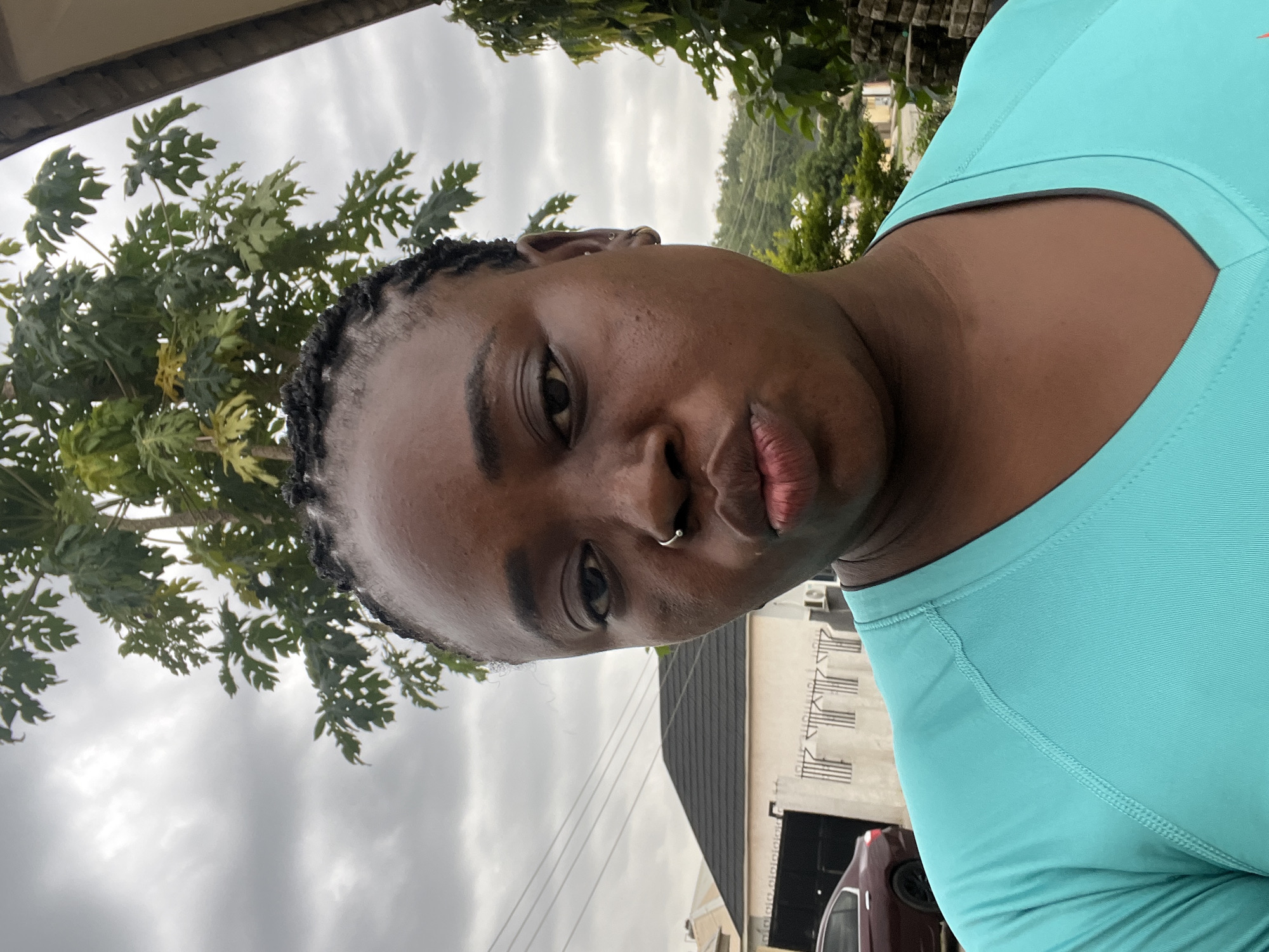 A close-up, selfie-style photo shows a woman with dark skin and braided hair. She is wearing a bright teal, V-neck, short-sleeved shirt. Her lips are pursed, and she has a silver hoop nose ring. In the background, a papaya tree with large green leaves is prominent against an overcast sky. To the left of the tree, a white building with a dark, peaked roof and a black garage door is visible, with a maroon car parked in front. Utility lines run horizontally across the sky. To the right of the papaya tree, a stone wall and more greenery are present. The overall scene appears to be an outdoor residential area in Abuja, Nigeria.
