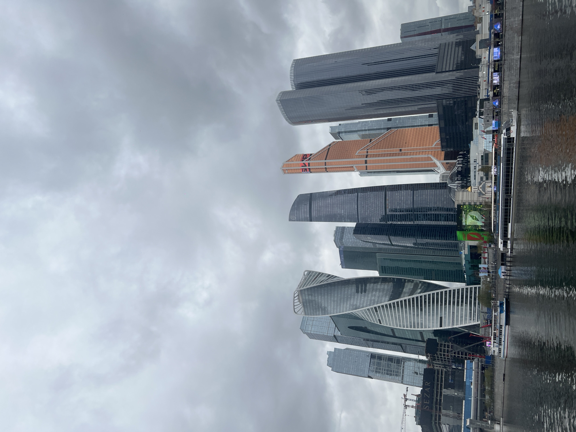 The image shows a skyline of modern skyscrapers in Moscow, Russia. The buildings are reflected in the water below. The sky is overcast and gray. There are several large buildings, including a distinctive twisting white skyscraper. There is a boat on the water and construction sites visible near the water.