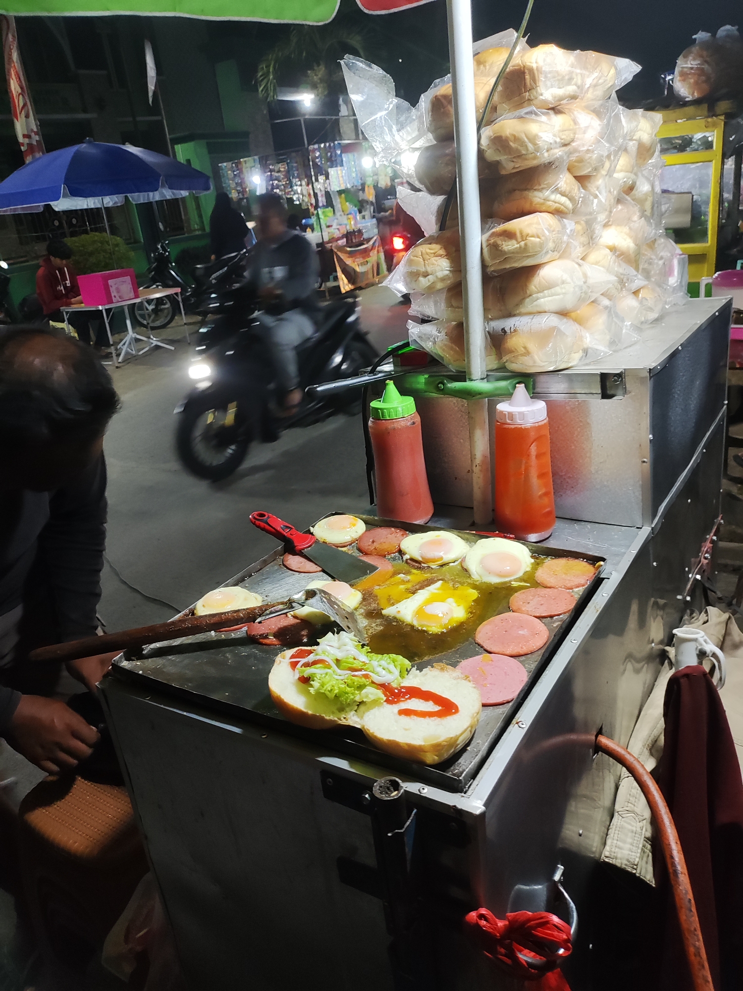An outdoor food stall is set up at night in Panongan, Indonesia, with a charcoal grill in the foreground where burgers are being prepared. Several burger buns are stacked on a cart, some are open and being assembled with lettuce and condiments. A person, partially visible, is operating the grill, using tongs and a spatula. A motorbike is seen passing by in the background, its headlights illuminating the scene. The stall has a canopy and appears to be situated on a street, with other stalls or shops visible in the distance.