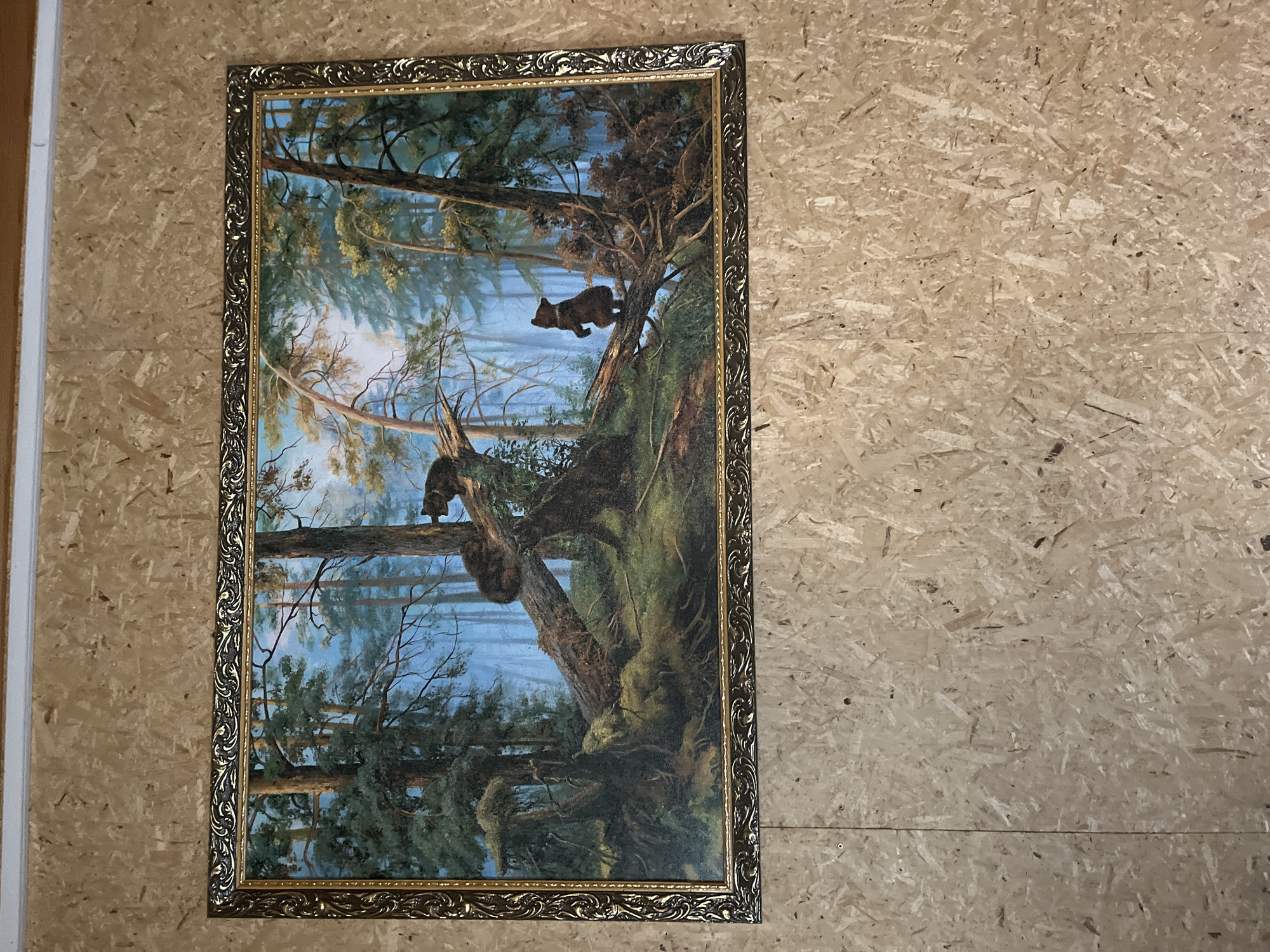 The image depicts a framed painting of a forest scene with two bears. The painting features tall evergreen trees, a fallen log covered in moss, and a misty blue background. One bear is perched on the fallen log, while another stands on its hind legs on a smaller branch. The painting is hung on a wall made of particleboard. The location of this image is not specified in the provided information and cannot be determined.