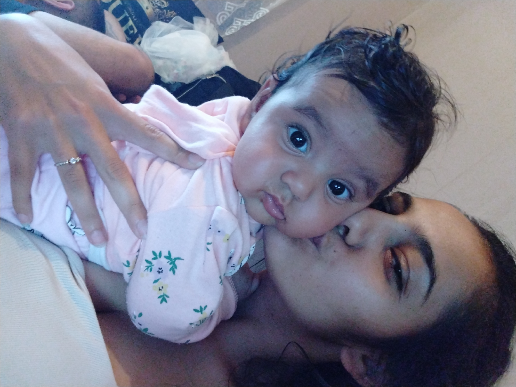 A photograph captures a woman lying down, kissing a baby in a pink floral-patterned outfit. The woman has dark hair and is lying on her side. The baby, positioned above her, looks directly at the camera. A hand with a ring on the ring finger is visible, supporting the baby. The location is Unknown, Unknown.