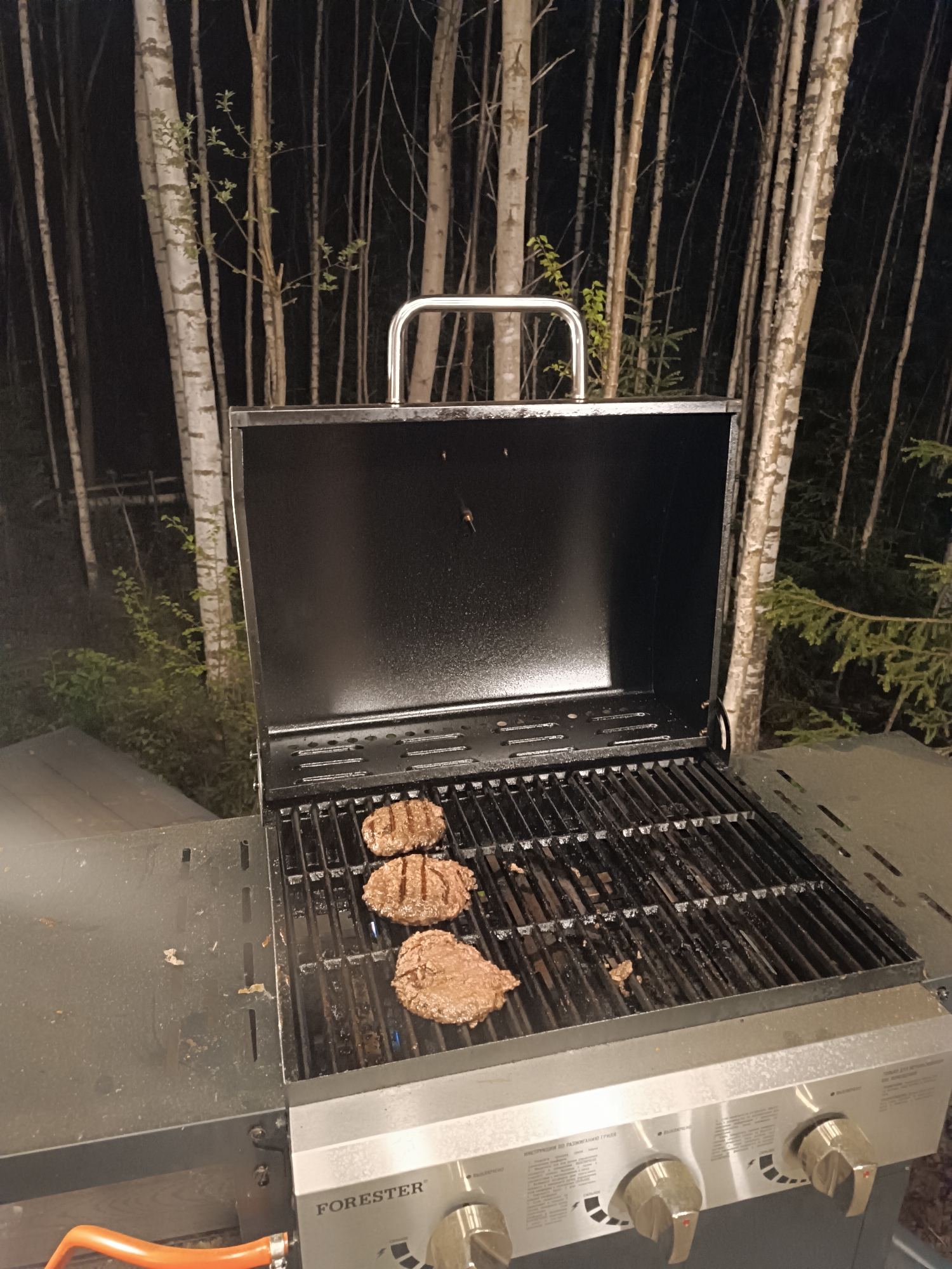 A grill sits on a flat surface, with three hamburger patties cooking on the grates. The grill is open, revealing a black interior. The grill has a silver control panel with three knobs. The grill brand is "FORESTER." An orange propane hose is visible. In the background, a stand of slender, light-colored trees extends into the dark. The location is Сепыч, Russia.