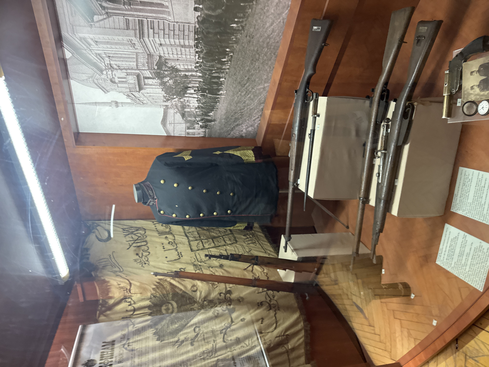 This image shows a display case in the city of Plovdiv, Bulgaria. The case contains several artifacts relating to the Ottoman Empire.
At the top of the case is a black and white photograph of a building, with a crowd of people outside. Below this is a blue military jacket. Beneath the jacket is a yellowed textile with Arabic writing and drawings on it. Under the textile is a sign.
To the right of these items are three rifles, mounted vertically. Next to the rifles is a shelf with more items on display, including a black leather belt. There are also signs in front of the rifles. The walls of the display case are made of wood. The display is inside, and the room is lit by a bright light.
