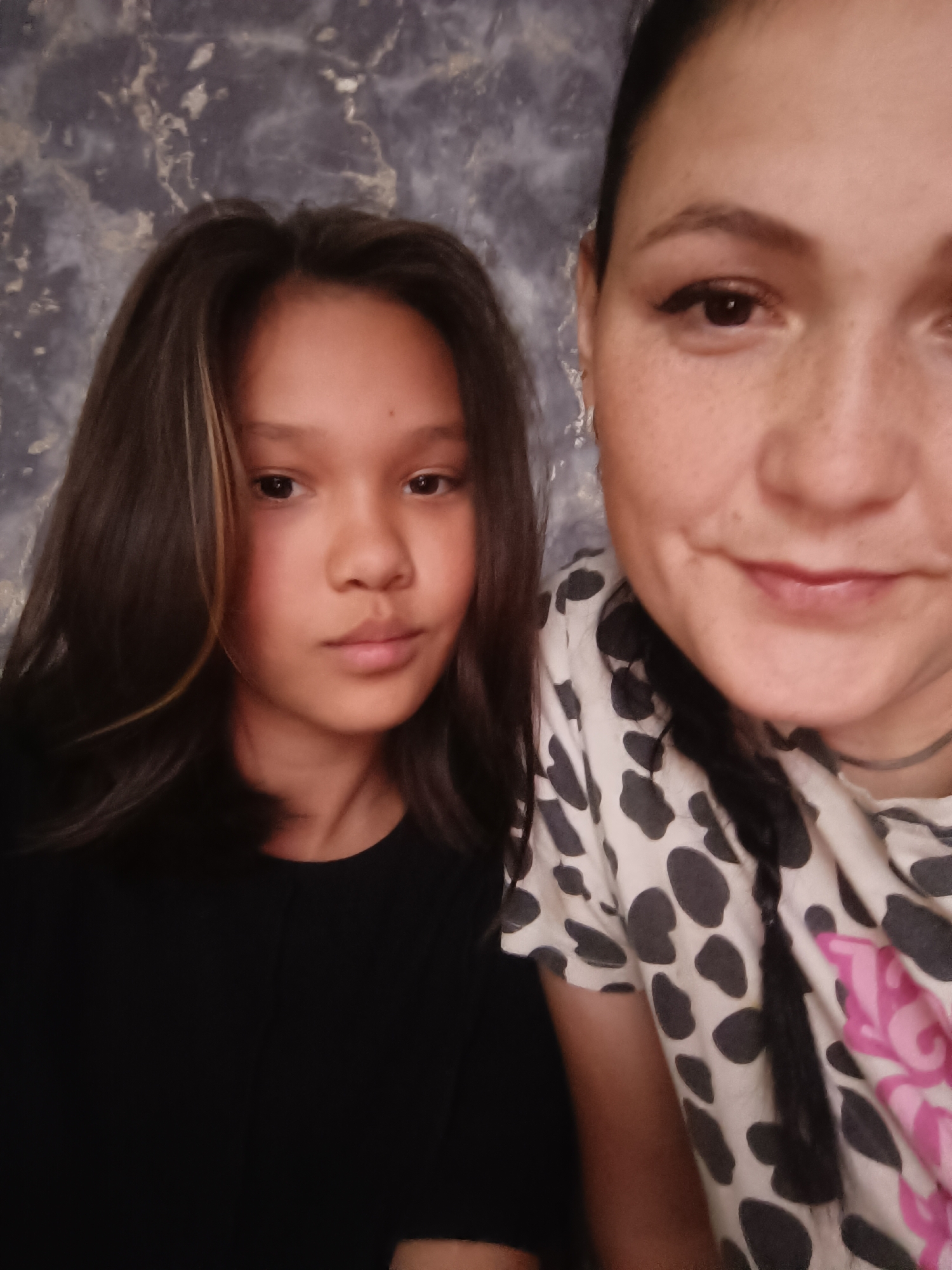 The image is a close-up selfie of two individuals, a young girl and an adult woman, positioned side-by-side. The background appears to be a wall with a marbled pattern in shades of gray and white.
The young girl is on the left side of the frame, facing the camera with a neutral expression. She has dark, shoulder-length hair with some lighter streaks and is wearing a black t-shirt.
The adult woman is on the right side of the frame, also facing the camera. She has her hair pulled back, and a braid is visible. She is wearing a white t-shirt with a black polka-dot pattern and some pink graphic design. She is smiling slightly. Both individuals appear to be indoors. There is no indication of the location being in the city of Karsha, Russia.