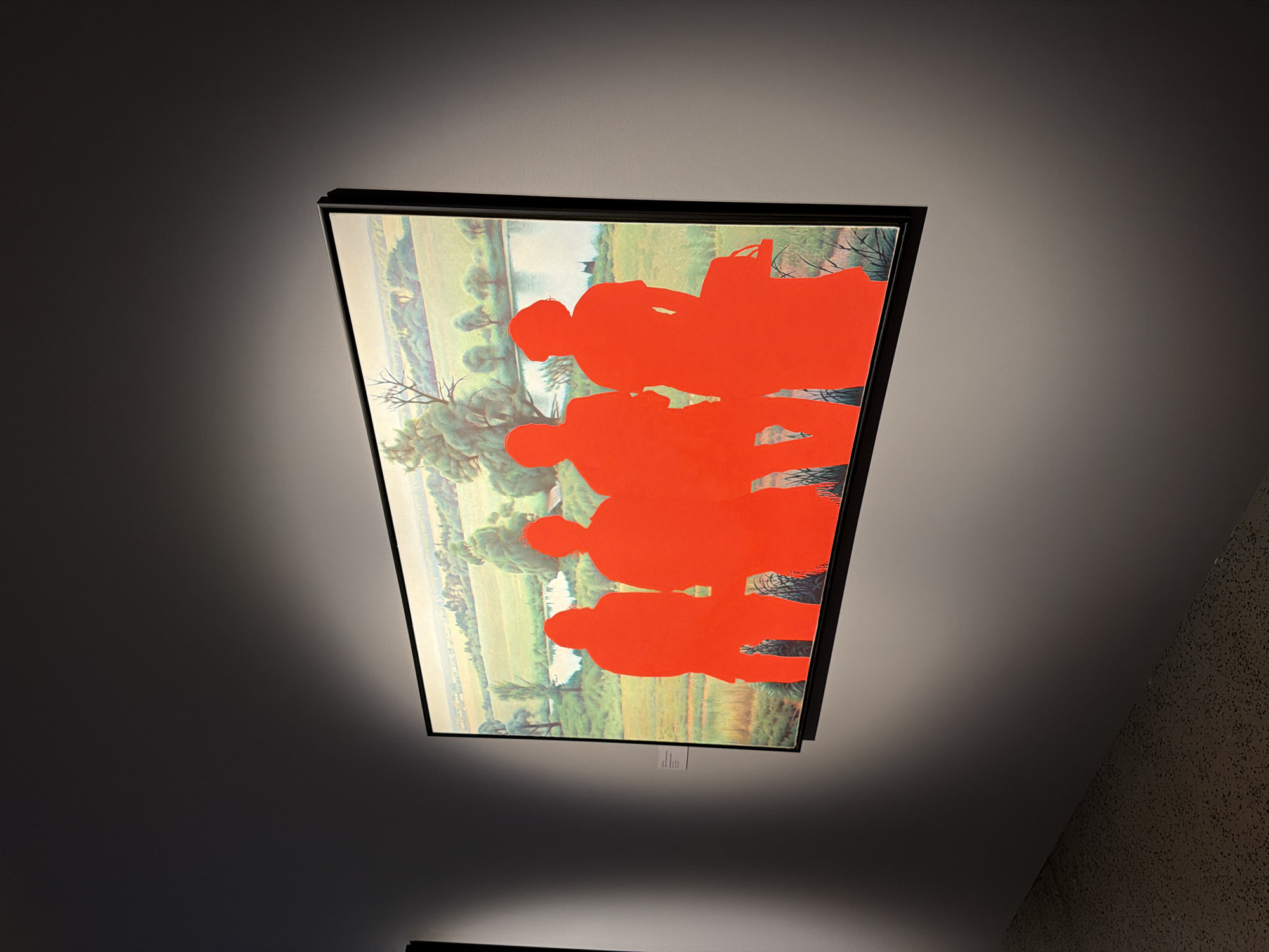 A framed artwork is displayed, tilted to the right, on a dark wall that transitions to a textured wall on the right. The artwork features a landscape with a body of water, lush green fields, and trees. Overlaying the landscape are bright red silhouettes of several figures. The silhouettes appear to be of people, possibly walking or standing together. The style of the artwork is a juxtaposition of a realistic landscape with abstract, solid color figures. The lighting casts a strong shadow from the artwork onto the wall, suggesting a directional light source from above and to the left. There is a small white label with black text visible on the bottom edge of the frame, but the text is not legible. The overall scene evokes a surreal or dreamlike quality due to the stark contrast between the realistic background and the abstract foreground figures.