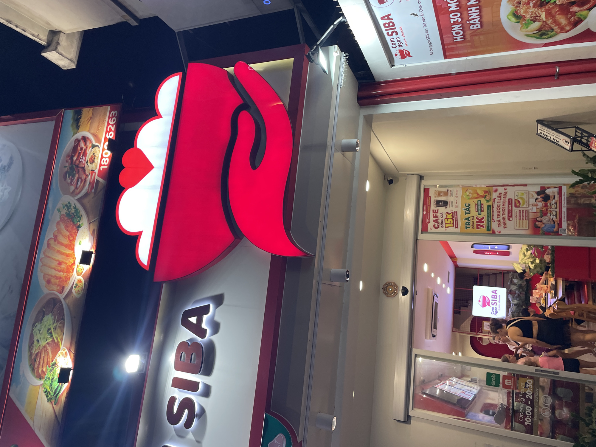 The image shows a street-level view of a restaurant named "SIBA" in Thủ Đức, Vietnam. The restaurant features a prominent red and white sign with the "SIBA" logo, which appears to be a stylized hand. The facade of the restaurant is brightly lit, suggesting it's either evening or the interior lighting is strong.
On the left side of the image, there are large display posters showcasing food items like pho and other dishes, with text including a phone number "180 6263". Above the restaurant entrance, a sign reads "Cơm SIBA Ngon" with the tagline "ĂN SẠCH - SỐNG KHỎE" (Eat Clean - Live Healthy). Further to the right, there are advertisements for "CAFE" for 15K and "TRÀ TẮC" (iced tea) for 7K, along with a promotion for paying in advance for all meals.
The entrance to the restaurant is visible, showing a clean, well-lit interior with tables and chairs. A few people are seen inside, and a woman is standing near the entrance, possibly a staff member or a customer. The overall atmosphere appears inviting and modern. The presence of a "Grab" logo near the door suggests that delivery services are available. The opening hours are displayed as "10:00 - 20:30, Monday - Sunday". The time of day is likely evening due to the artificial lighting and dark sky.