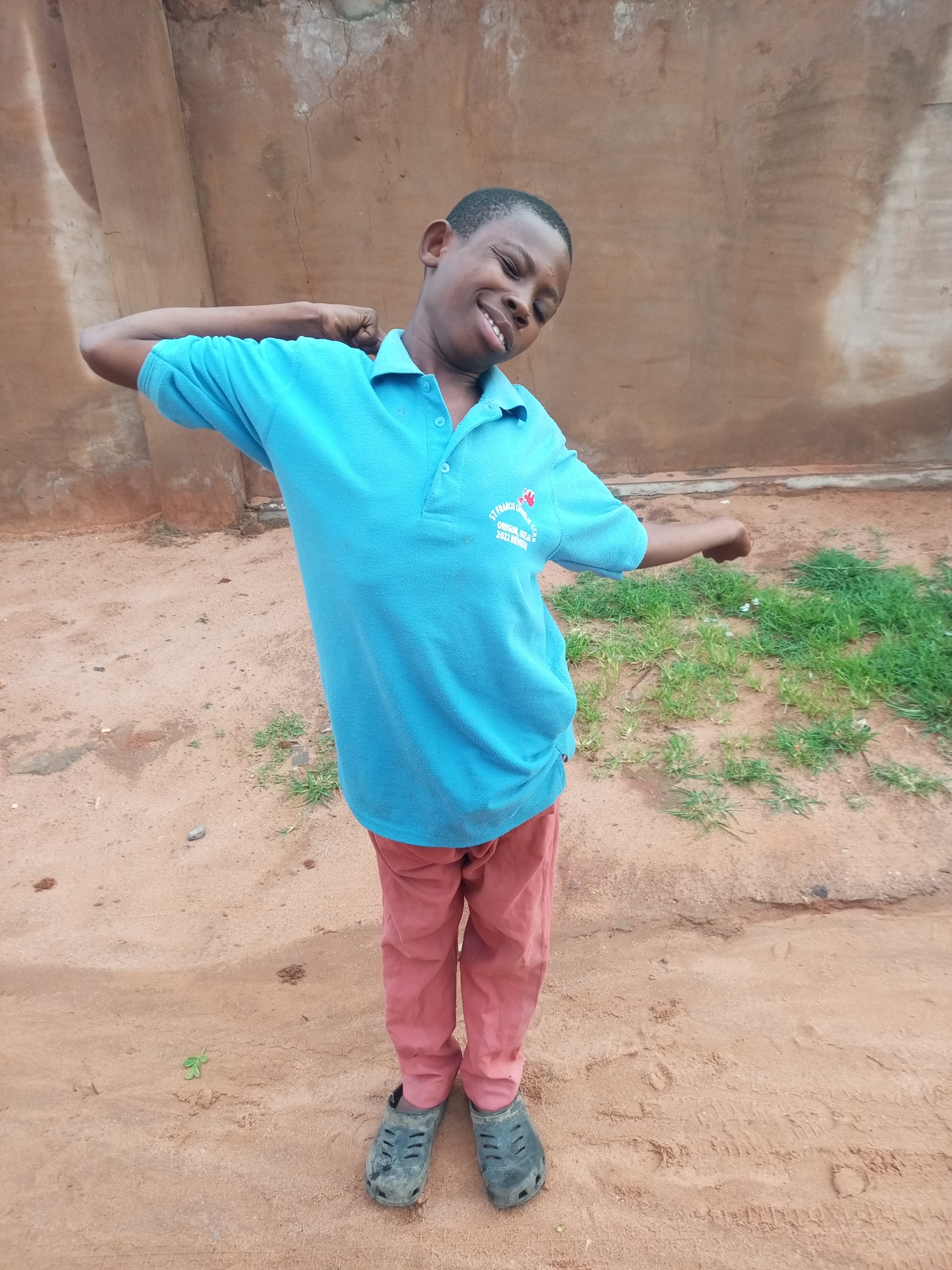 A young boy with dark skin and short hair is standing in an outdoor setting, facing slightly to the right of the camera. He is wearing a bright blue polo shirt with a logo on the left chest and coral-colored pants. His left arm is raised and bent at the elbow, with his fist clenched, and his head is tilted back, his eyes closed, and his mouth open in a wide smile. His right arm is also raised and bent at the elbow, with his fist extended. He is wearing dark, slip-on shoes. The background consists of a rough, textured, reddish-brown wall and a sandy ground with patches of green grass. The overall impression is one of youthful exuberance and playfulness. The location is identified as a school in Tempure, Nigeria, indicated by the logo on the boy's shirt, which reads "ST. FRANCIS CATHOLIC SEC SCH, OREGUN, IKEJA, 2022 RURAL".