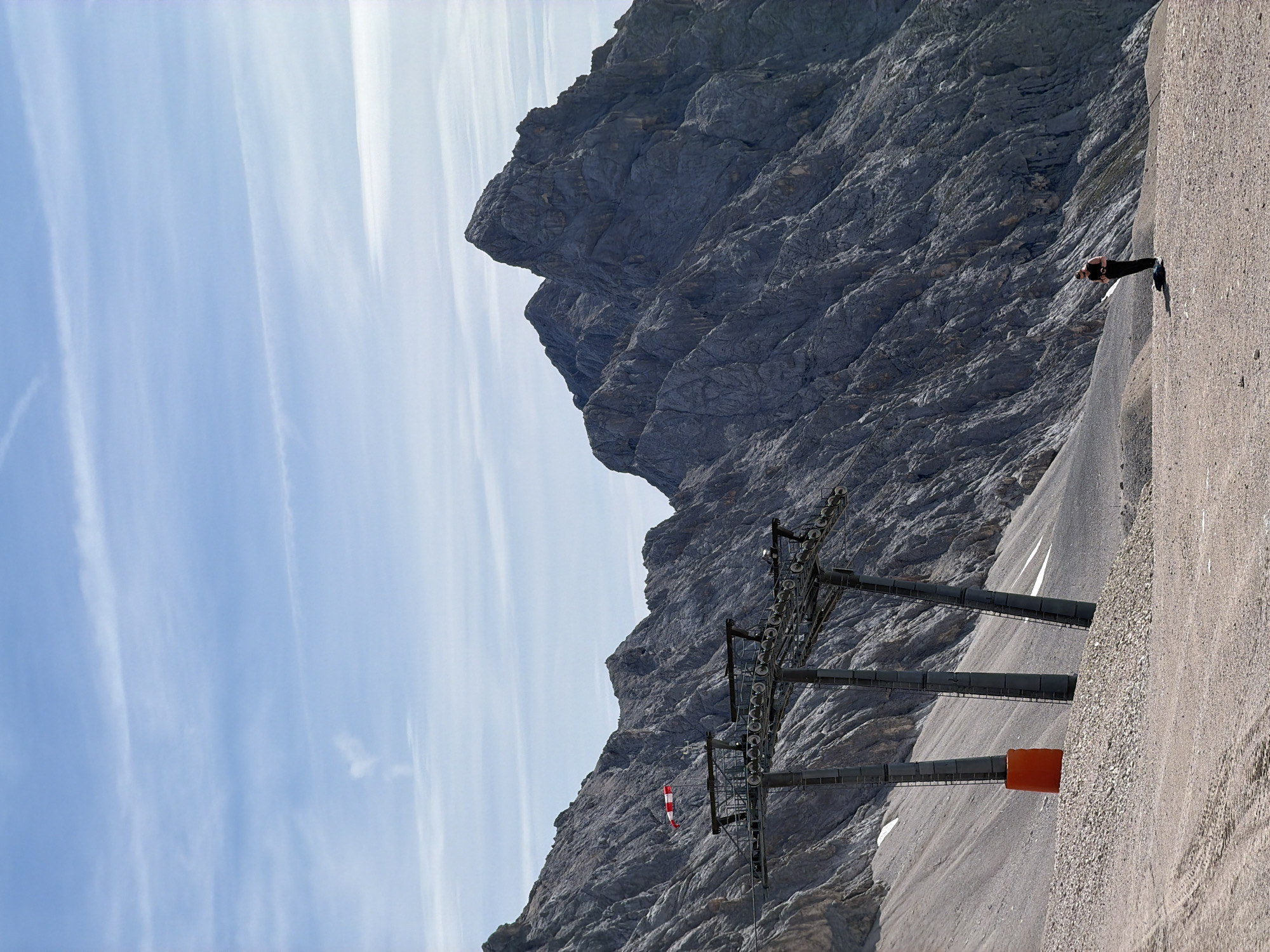The image depicts a mountainous landscape with a ski lift in the foreground and a person standing on a gravel path in the background. The ski lift towers are grey, with an orange base on one of them. A red and white flag is attached to the ski lift structure. The mountains are steep and rocky, with some snow patches visible on their slopes. The sky is blue with scattered white clouds. The person in the background is wearing dark clothing and appears to be holding something in their hands. The scene is set in a high-altitude, rugged environment. This location is likely the Zugspitze, Germany's highest peak, which is accessible from Garmisch-Partenkirchen.