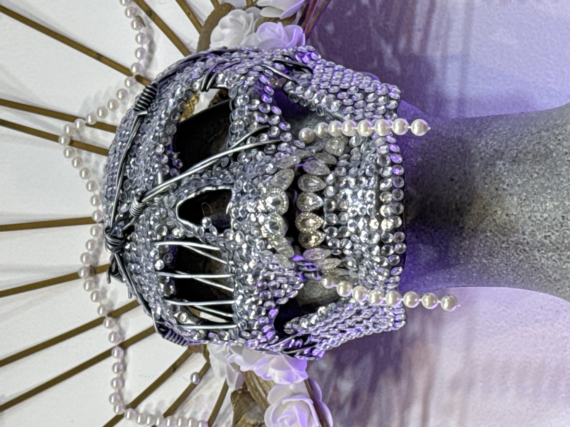 The image shows an elaborate skull sculpture. The skull is adorned with numerous small, sparkling rhinestones and embellished with pearl strands. It is attached to what appears to be a vase. The skull is also framed by a white parasol decorated with pearl strands and white roses. The image was most likely taken indoors under purple lighting. There is no discernible information to determine if the image was taken in Sofia, Bulgaria.