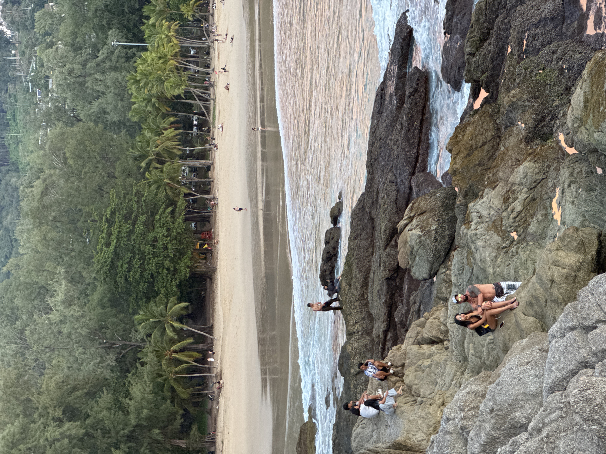 This image captures a scene at Bang Tao Beach in Choeng Thale, Thailand, a popular tourist destination known for its long stretch of sand and coastal amenities. In the foreground, a rocky outcrop lines the shoreline. On these rocks, several groups of people are visible. Closest to the viewer, a couple sits on a large boulder; the woman is wearing a black bikini top and shorts and sunglasses, while the man is shirtless, wearing plaid shorts and a white baseball cap. Further up the rocks, to the left, a group of four young people are seated, appearing to be resting or conversing. To the right, a man stands shirtless near the water's edge, looking out at the sea.
Beyond the rocky area, the sandy beach stretches out, lined with numerous palm trees and other vegetation, forming a lush backdrop. Several people are scattered across the beach, some walking, others relaxing on the sand. The water is calm with gentle waves lapping at the shore. The overall atmosphere suggests a relaxed, late afternoon or early evening scene at a tropical beach.