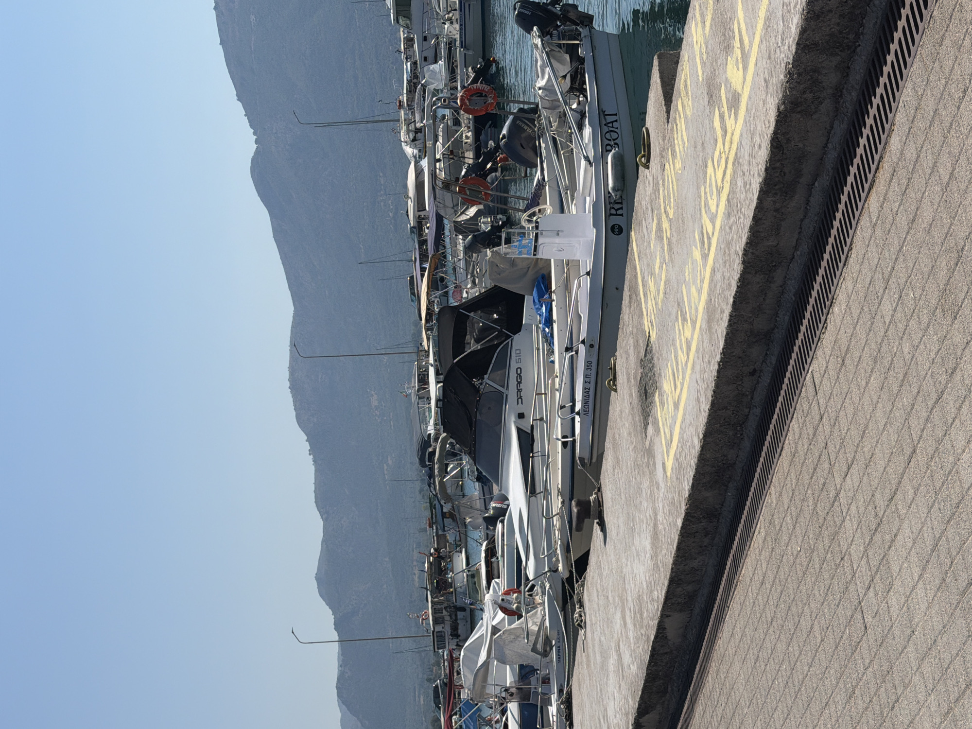 The image depicts a sunny day at a marina in the Municipal Unit of Alyzia, Greece. The foreground shows a concrete pier with yellow painted lines and a grated drain. Several boats are docked in the water. The closest boat in the foreground has "RED BOAT" written on its side in black lettering. Other boats are visible behind it, including some with canopies and outboard motors. In the background, a range of blue-hued mountains under a clear sky forms the horizon. The water is calm and reflects the blue of the sky.