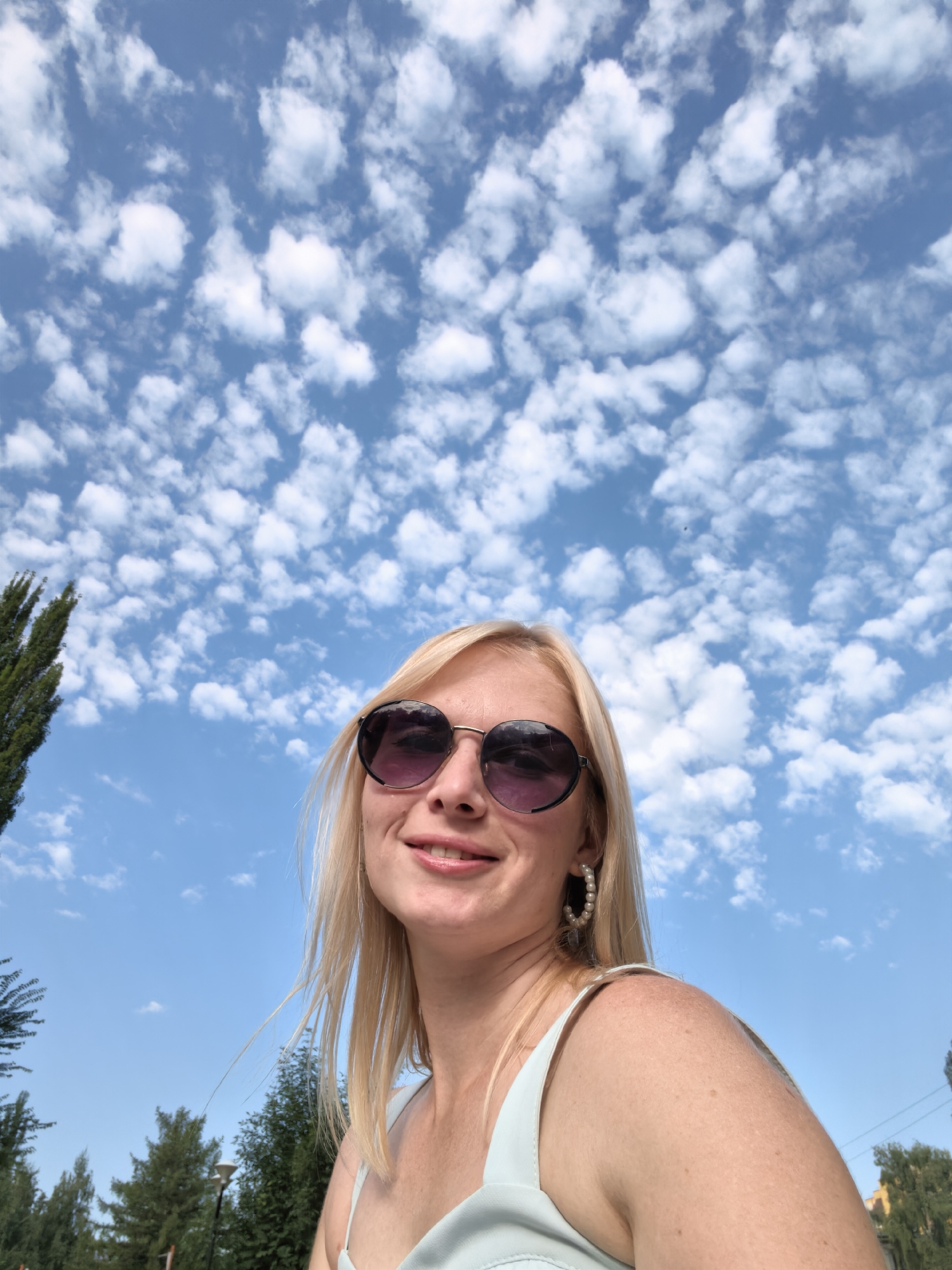 A blonde woman wearing sunglasses and pearl earrings smiles as she looks up at a cloudy blue sky. She is outdoors, with trees and a lamppost visible in the background. The image is a selfie, taken from a low angle. The location is not specified in the image or any associated metadata.