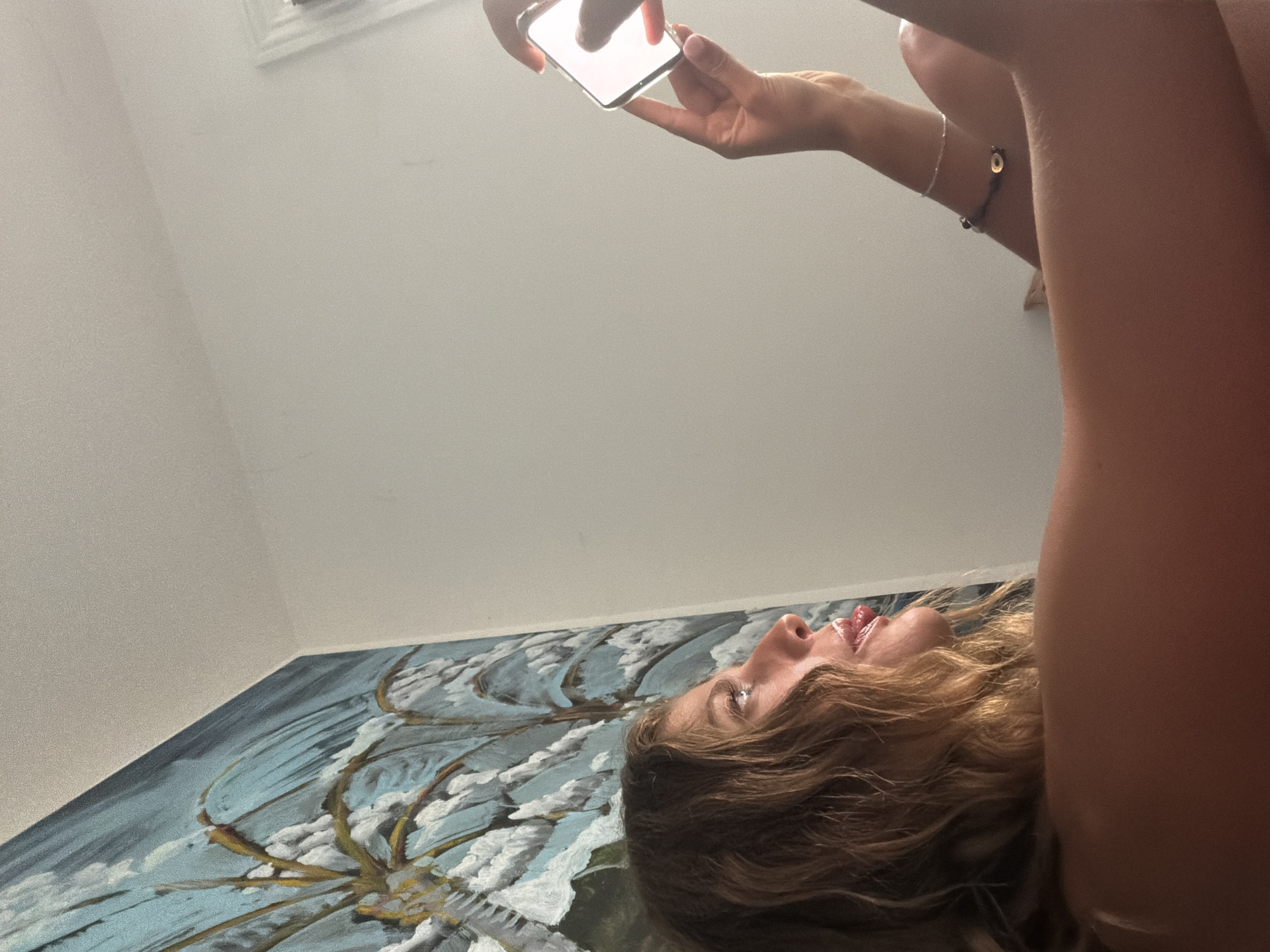 A person lies on a painting, looking up and holding a phone. The location appears to be indoors, but there is no discernible indication of Malibu or the United States of America.