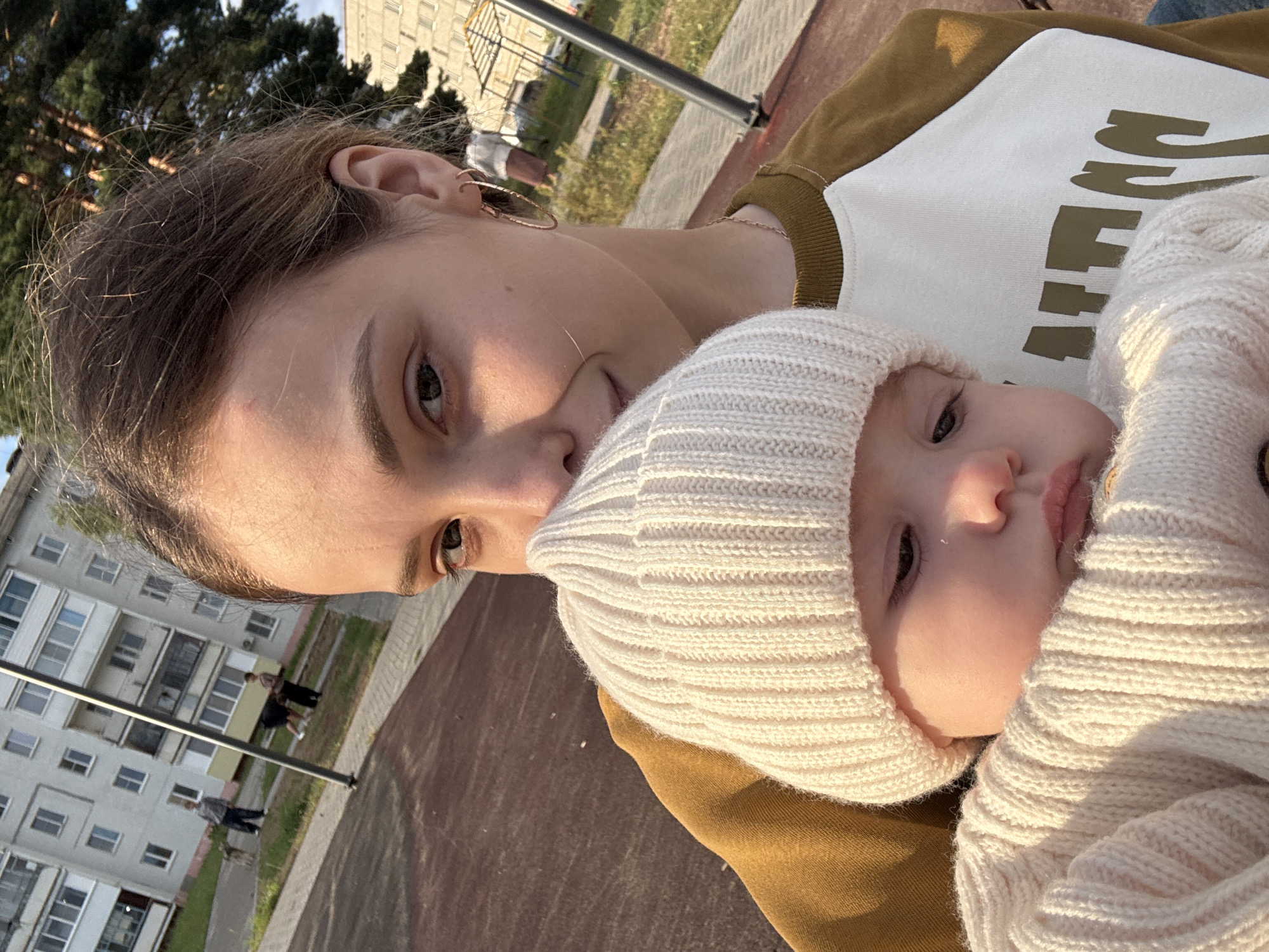 A young woman holds a baby outdoors in Noviy, Russia, on a playground. The woman, with brown hair pulled back and wearing hoop earrings, is holding the baby close to her chest. The baby, wearing a white knit hat and sweater, looks down with a neutral expression. Behind them, a modern apartment building and playground equipment are visible, suggesting a residential area. The lighting indicates it is late afternoon or early evening.