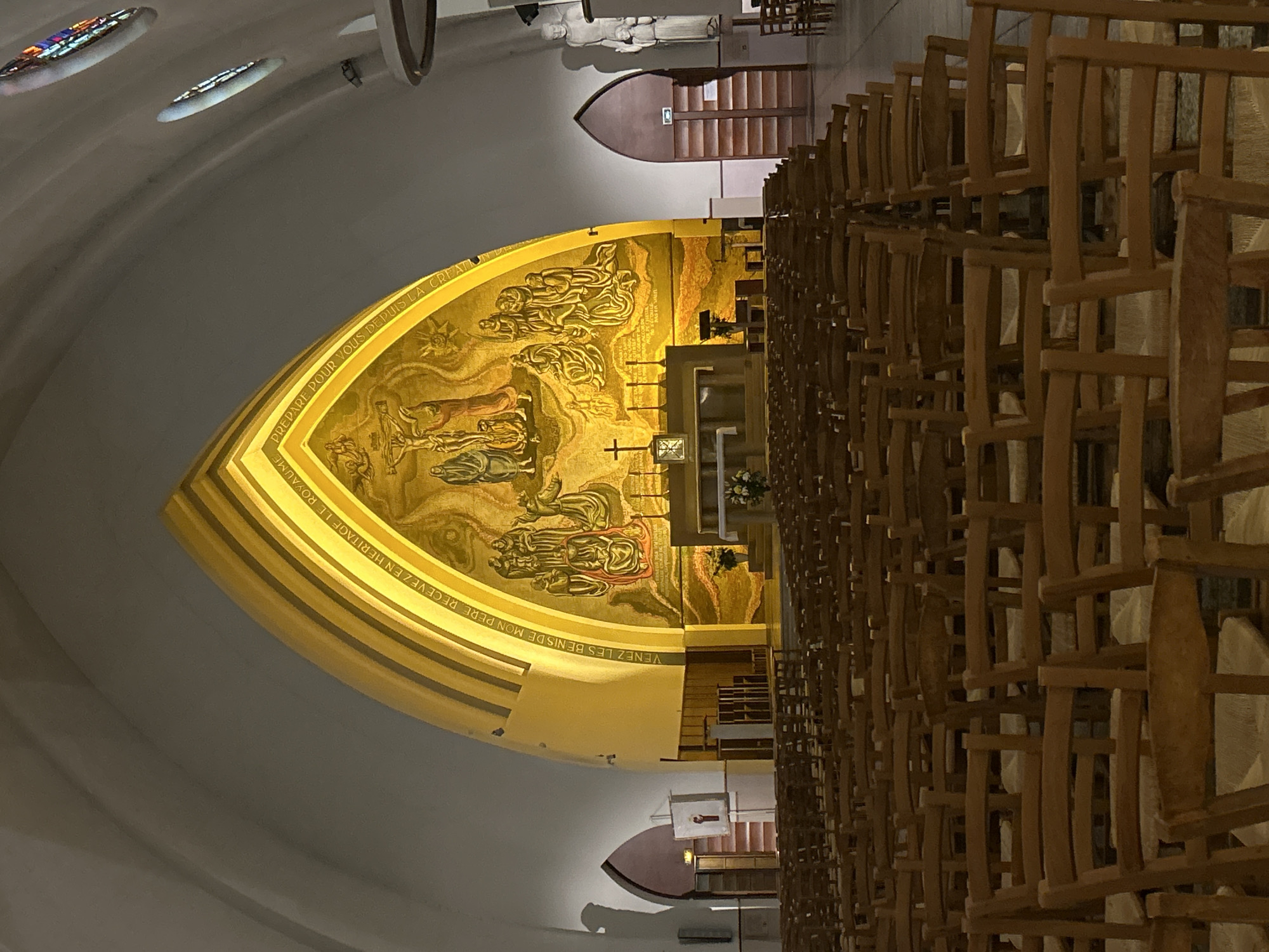 This interior shot, rotated 90 degrees clockwise, depicts what appears to be a church or chapel. The perspective is from the side, looking towards the altar area. A large, curved, golden mosaic dominates the focal point, depicting numerous figures and scenes, including a depiction of Jesus on the cross and other individuals, possibly saints or celestial beings. This mosaic is brightly illuminated, casting a warm glow.
Below the mosaic, an inscription in French is visible. Portions of the text read: "PREPARE POUR VOUS DEPUIS LA CREATION" and "VENEZ LES BENIS DE MON PERE. RECEVEZ EN HERITAGE LE ROYAUME," translating to "Prepared for you since creation" and "Come, blessed of my Father. Receive the kingdom as your inheritance," likely a biblical quote (Matthew 25:34).
To the right, rows of simple wooden chairs or pews are arranged, some appearing stacked, suggesting a flexible seating arrangement or storage. In the upper background, a white statue of a robed figure stands near a simple, dark wooden door or structure. The ceiling is a large, light-colored arch with several small, oval stained-glass windows that admit muted light. The overall lighting is dim and atmospheric, emphasizing the illuminated mosaic and creating a serene ambiance within the religious space. No specific activity is taking place; it's a static view of the building's interior.