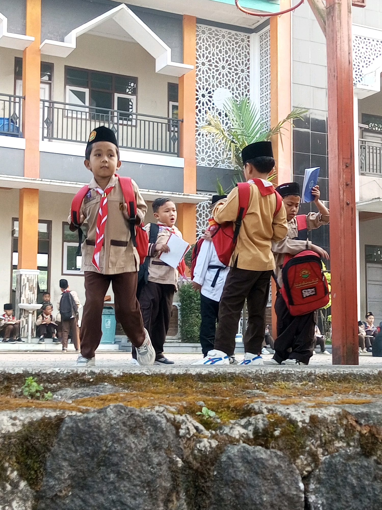 In Kalibeber, Indonesia, a group of children wearing scout uniforms are gathered outside a building. The central figure, a young boy in a tan uniform with a red and white scarf, stands prominently, facing forward with a red backpack. To his right, another boy in a similar uniform holds a blue book above his head. Other children are visible in the background, some seated and others standing, also in uniform. They are positioned in front of a multi-story building with orange and gray accents, featuring decorative white grilles on its facade. The ground in the foreground is covered with moss-covered rocks and sparse vegetation.