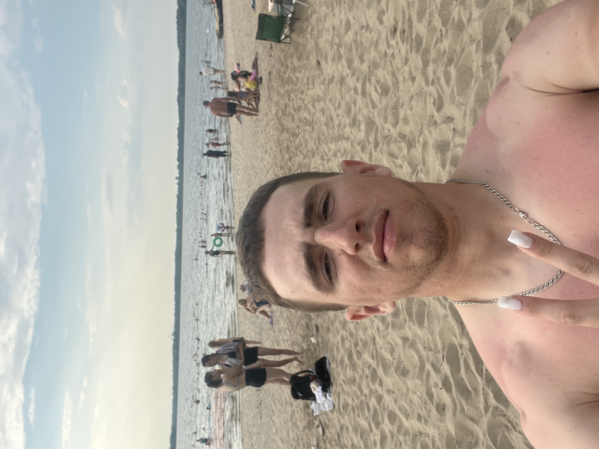 This is a selfie taken at a beach on a bright day, likely in the afternoon, judging by the lighting. The foreground is dominated by the upper body of a young man, facing the camera. He has short brown hair and is wearing a silver chain necklace. His skin appears tanned and slightly sunburnt on his shoulder and chest. Beside him, a hand with white nail polish is giving a peace sign.
The background depicts a crowded beach scene. Numerous people are visible, some wading in the shallow water, while others are relaxing on the sand. The water is calm and appears to be a lake or a large river. There are several beach chairs and umbrellas scattered across the sand, indicating a popular recreational area. The overall atmosphere seems relaxed and summery, with people enjoying the warm weather. The terrain is sandy, typical of a beach. No text is visible in the image.