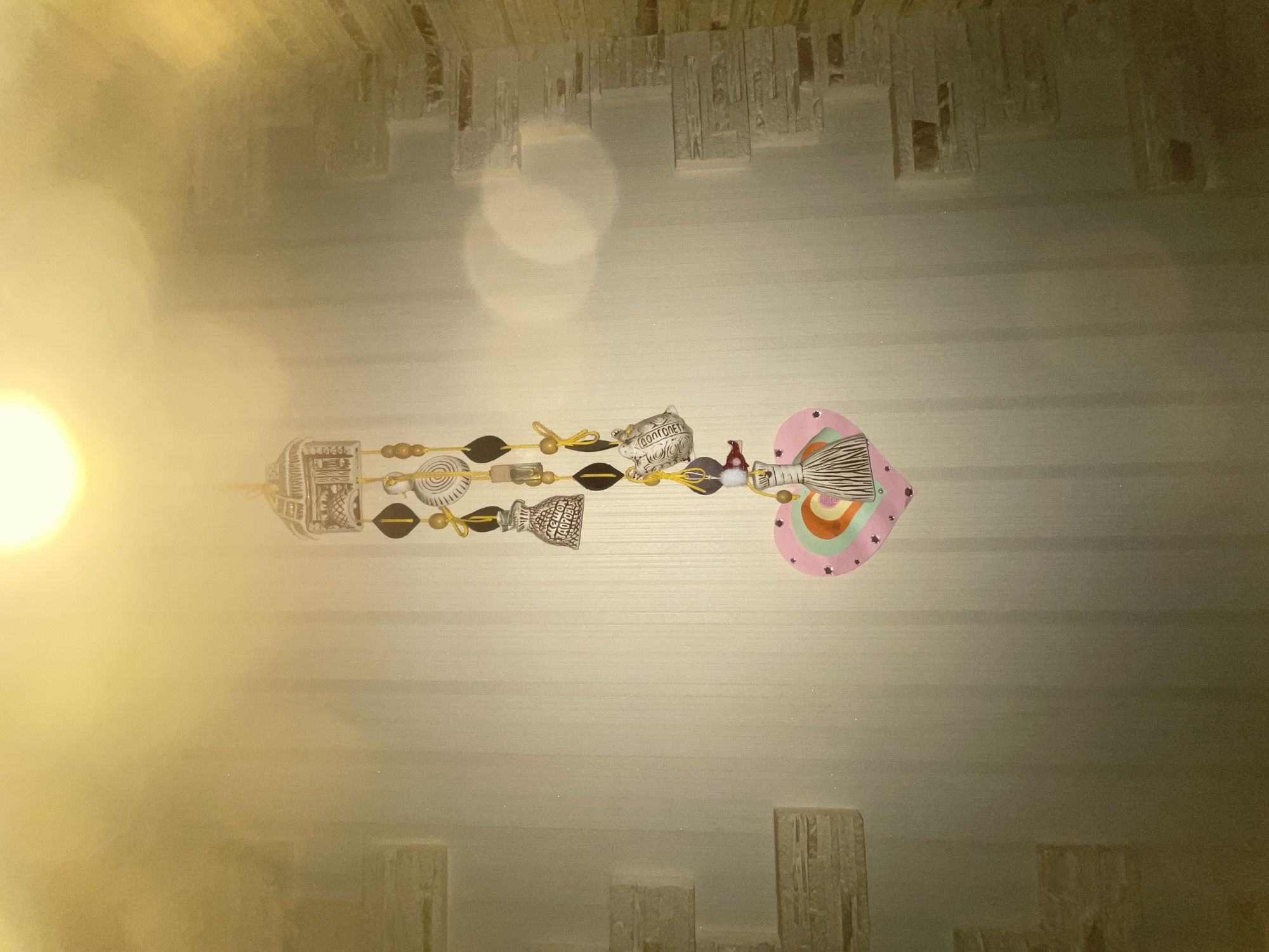 A decorative wall hanging featuring a heart-shaped cutout with colorful concentric circles and a tassel hangs on a beige, vertical-striped wallpapered wall. The hanging is adorned with various charms, including small bells and oval shapes. A bright light source illuminates the scene from the top, creating a soft glow. To the right of the hanging, a section of the wall is decorated with faux stone bricks. The overall lighting suggests an indoor setting, possibly a room. The location is identified as Unknown, Unknown.