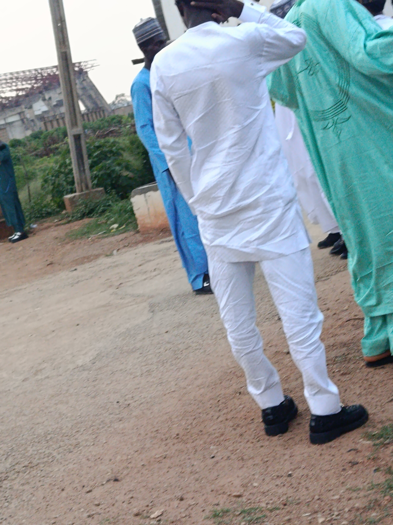 In Jos, Nigeria, a group of men, some wearing traditional white attire, stand near a construction site. The man in the foreground is dressed in a white tunic and pants with black shoes. To his right, another man wears a light green robe with intricate patterns and black shoes. Further in the background, a man in a blue tunic and cap is visible. The environment consists of a dirt road with overgrown grass and what appears to be an incomplete building structure behind the men. The sky is overcast, indicating a cloudy day.