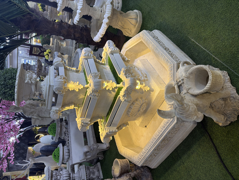 A three-tiered fountain with a goat head spilling into the bottom basin is the focal point. The fountain features intricate decorative details, including floral carvings and ornate edges. It's surrounded by other garden decorations and greenery, and the surface beneath is artificial grass. The overall scene is brightly lit, highlighting the texture of the fountain and nearby decor.