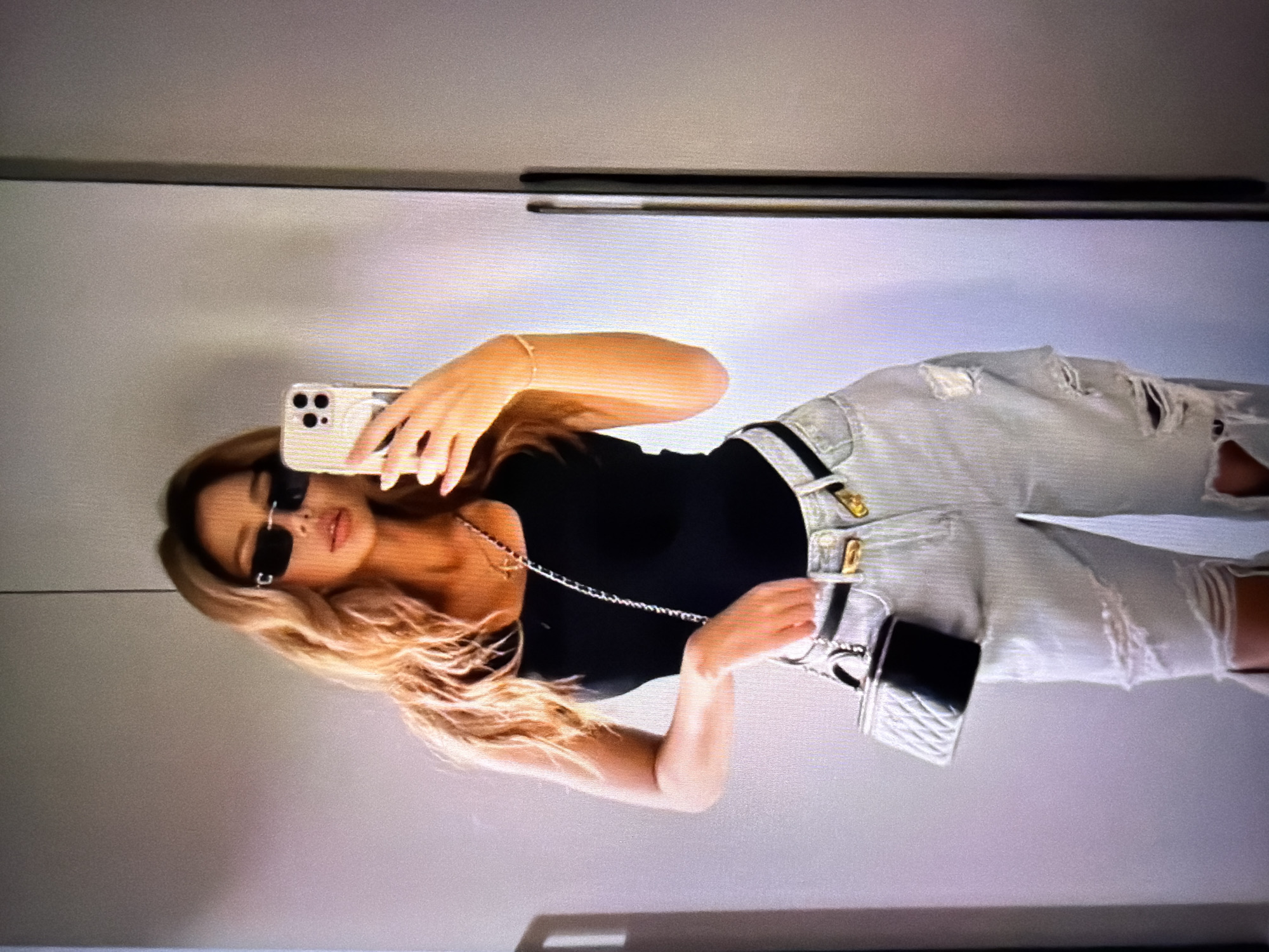 The image shows a woman taking a mirror selfie in what appears to be a domestic setting, possibly a bedroom or dressing room. She is wearing a black tank top, light-wash distressed jeans, a black belt with gold buckle accents, and black sunglasses. She is accessorizing with a small, quilted black and silver purse attached to her belt, a delicate gold necklace, and a bracelet. Her hair is long and wavy, with blonde and brown tones. She is holding a smartphone in her right hand, capturing the reflection. The background is a plain, light-colored wall, with a tall, narrow closet door visible on the right. There is no indication of the city of Beșalma, Moldova, or any other specific location.