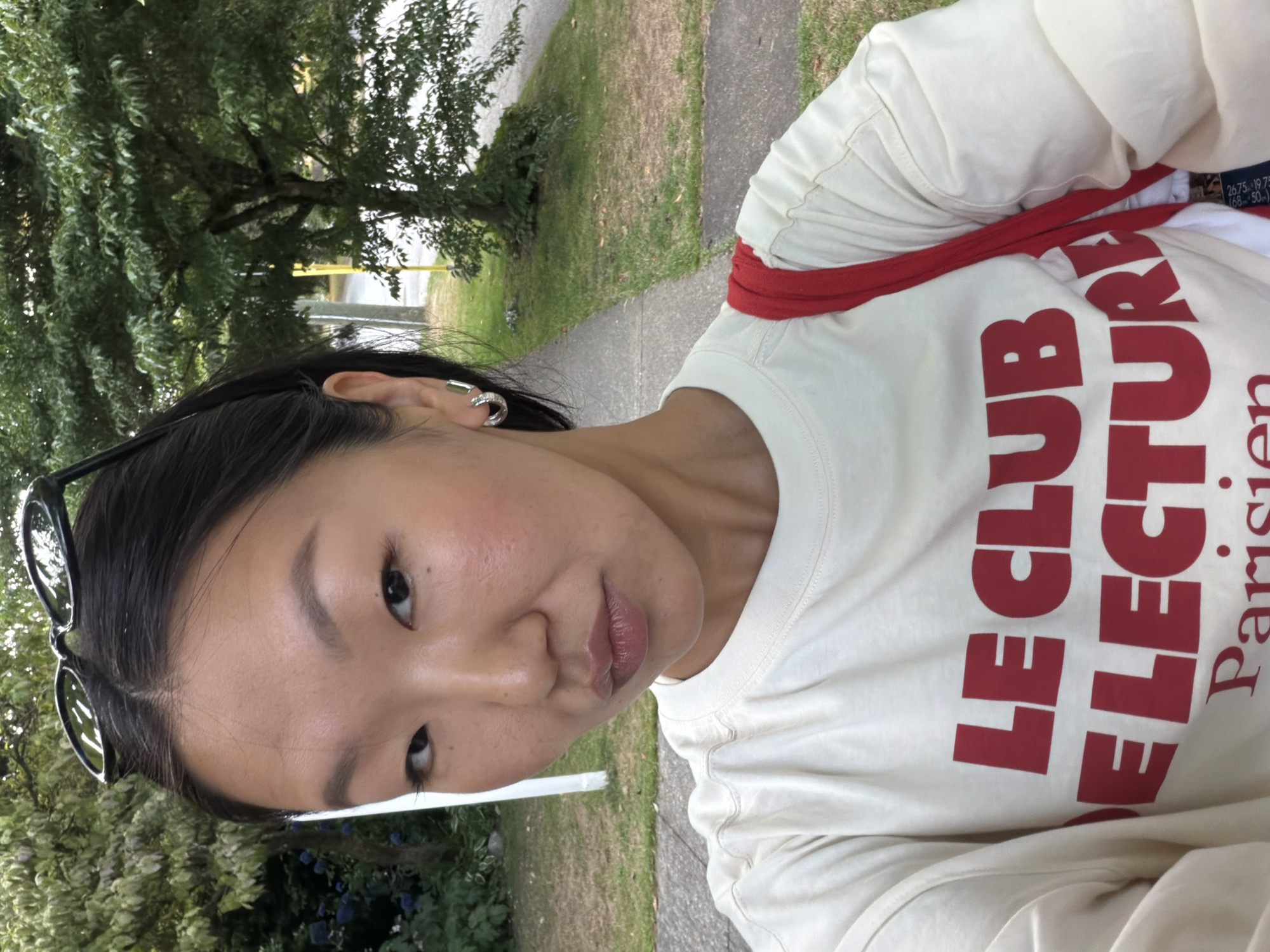 The image shows a close-up selfie of an East Asian woman outdoors, with a paved sidewalk and green foliage in the background. She has dark hair pulled back and is wearing large black sunglasses on top of her head, a cream-colored sweatshirt with red lettering that reads "LE CLUB DE LECTURE Parisien," and a red bag strap across her shoulder. She is also wearing silver hoop earrings. The lighting suggests it is daytime, possibly overcast. The location is identified as New Westminster, Canada, though specific landmarks are not visible.
