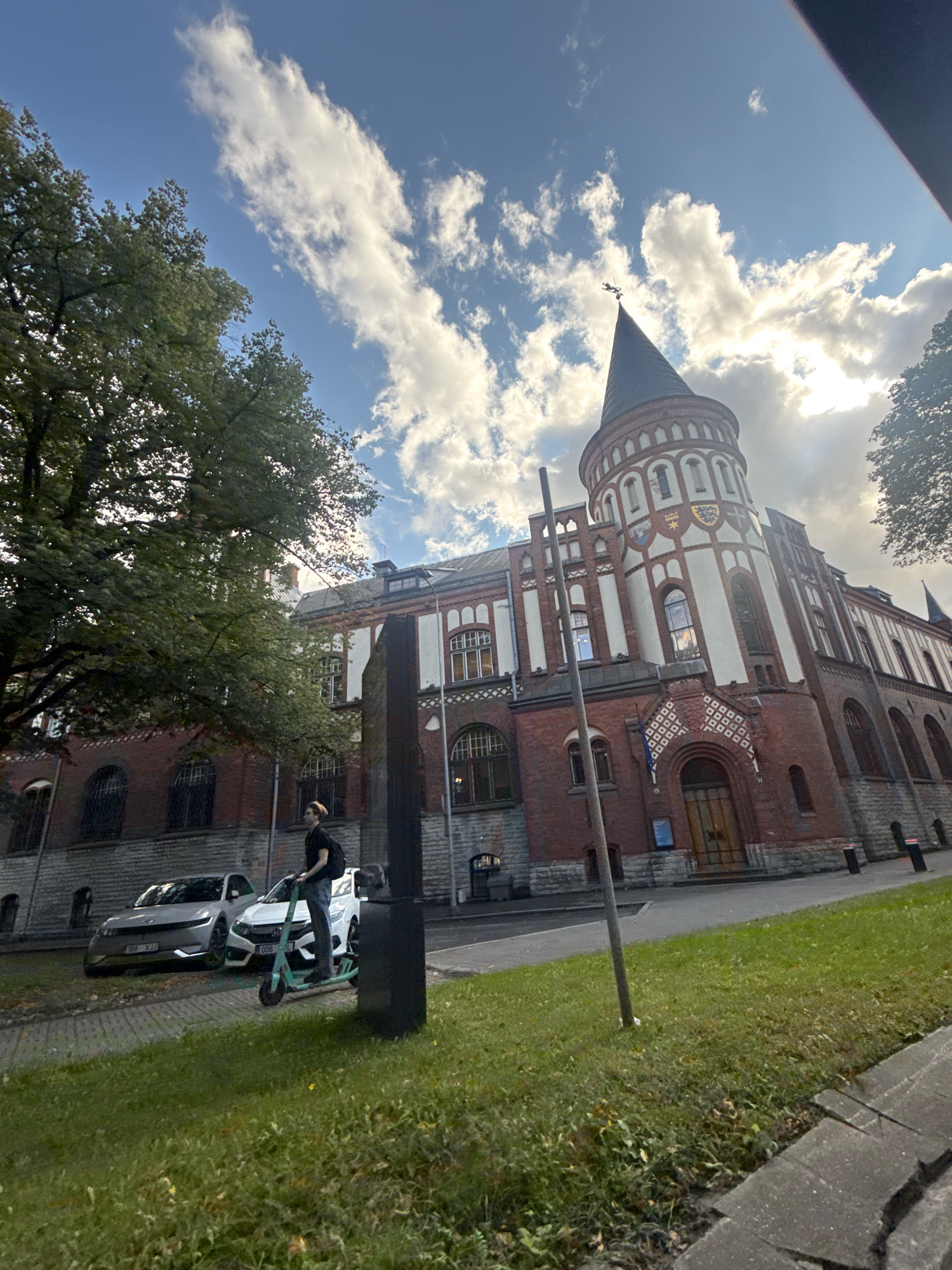 The image depicts an exterior view of a historic brick building with white trim and a conical tower, likely located in Tallinn, Estonia, given its architectural style. The building has arched windows and decorative elements. In the foreground, a person is standing on a green electric scooter. Two cars are parked to the left of the scooter. A large tree with green foliage is visible on the left side of the frame. The sky is blue with scattered white clouds, and the sun appears to be shining. The scene is set on a paved area adjacent to a grassy lawn.