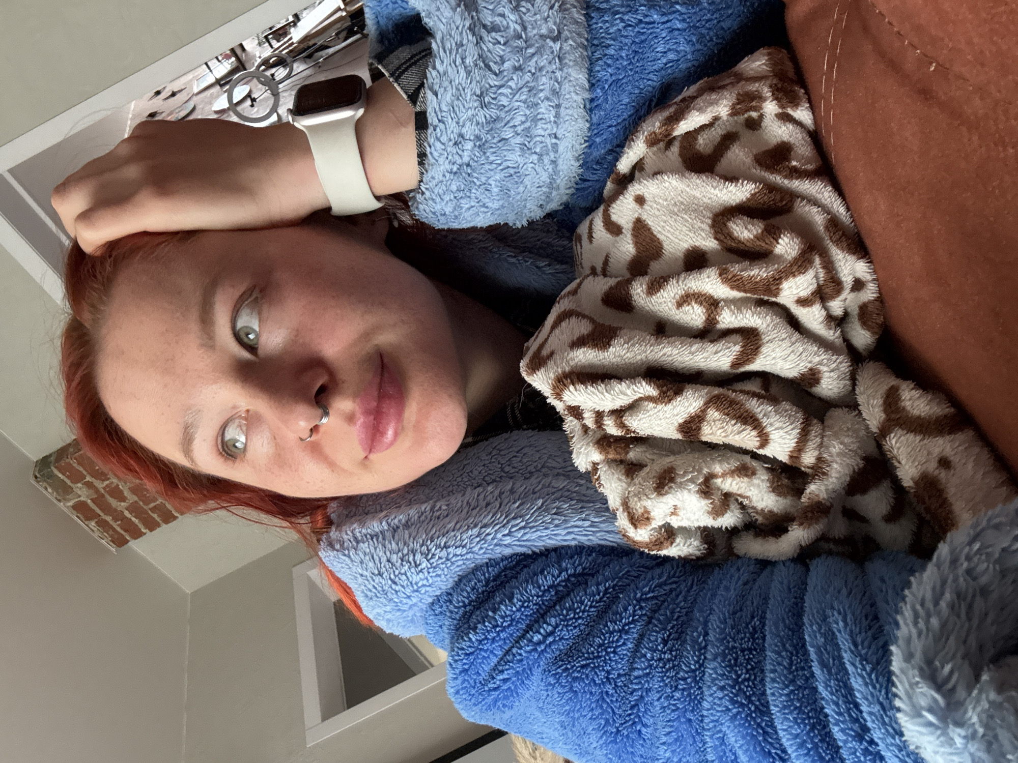 The media file displays a close-up, vertically oriented portrait of a person with auburn hair and light eyes. They are seen from a low angle, appearing to recline or lounge indoors. The person has a silver nose ring and a white Apple Watch on their left wrist. Their left hand rests gently on their head as they look directly at the camera with a relaxed expression and a slight smile.
They are wearing a blue fluffy garment, resembling a robe or a thick blanket, over what appears to be a plaid shirt sleeve. A second patterned blanket, featuring a brown and white abstract or animal print, is draped nearby. Next to the blanket, an orange-brown cushion or piece of furniture is visible.
The background shows elements of an indoor setting, including a white ceiling and wall, with a section of exposed red brickwork visible in a corner. Above the person's head, blurred circular objects or reflections can be seen. The overall impression is one of comfort and coziness. No visible text is present in the image. The location context for this image is Rostov-on-Don, Russia.