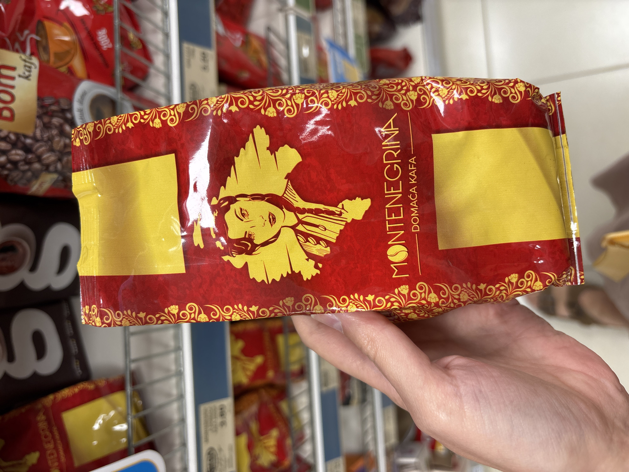 The image shows a hand holding a red and yellow coffee bag labeled "MONTENEGRINA DOMAĆA KAFA". The bag is adorned with a yellow illustration of a woman's profile and intricate golden patterns along the edges. The coffee bag is positioned in front of a shelf stocked with other coffee bags, suggesting a retail or supermarket setting. In the background, shelves are visible with various coffee packaging, and some blurry text indicating prices or product names. The scene appears to be inside a store, possibly a supermarket or a specialty shop, given the arrangement of products on shelves. The lighting is artificial, typical of an indoor retail environment. The photograph is taken from a slightly downward angle, focusing on the coffee bag in the foreground. No specific landmarks or details indicating the city of Boreti, Montenegro, are present in the image.