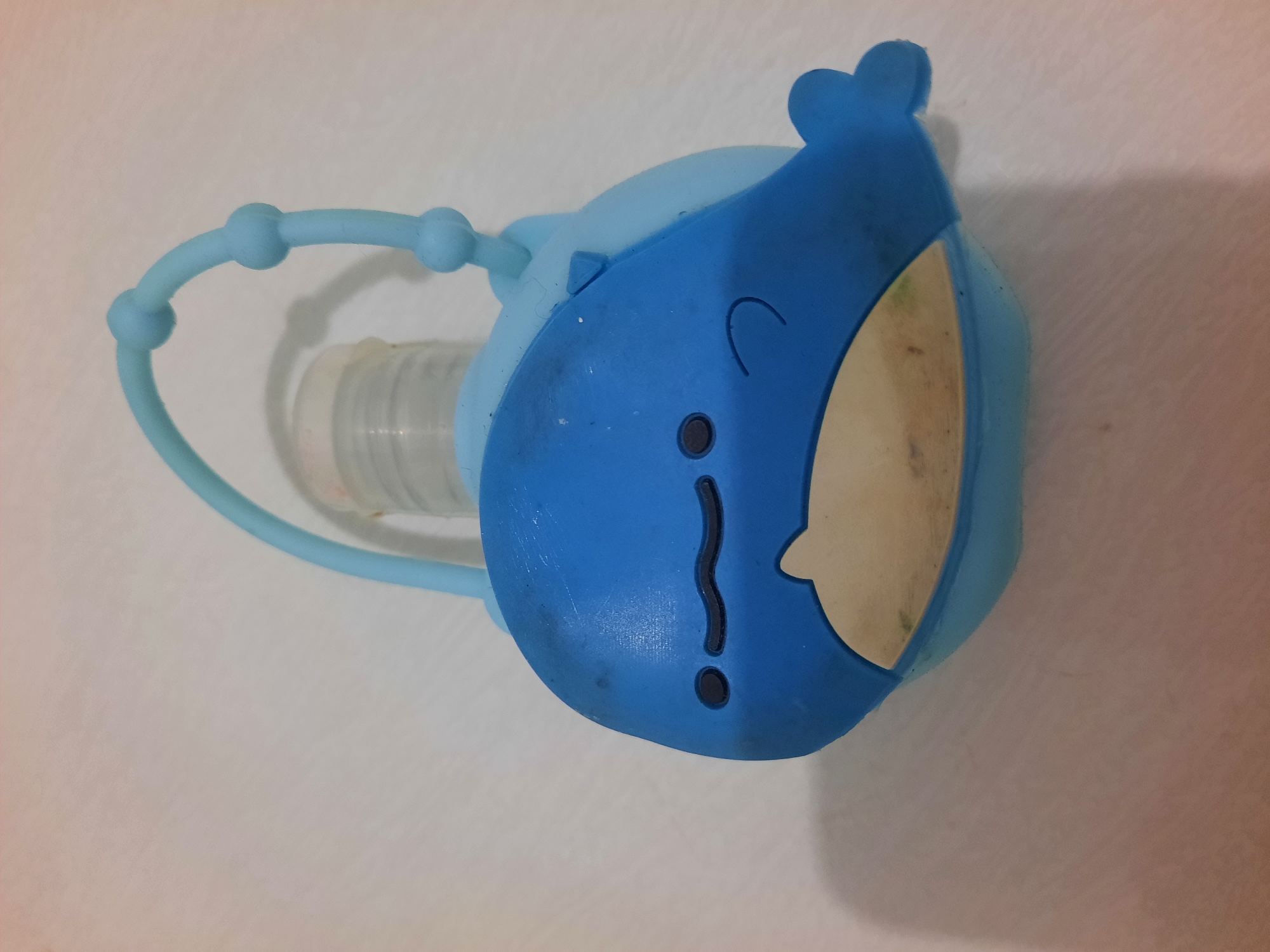 A blue silicone whale-shaped holder with a clear bottle inside is shown on a textured white surface. The whale has black eyes and a wavy black mouth. A light blue silicone loop with three small spheres is attached to the top of the whale, presumably for hanging. The bottle appears to be a hand sanitizer dispenser. There is some dust or debris on the whale's surface and inside the bottle's opening.