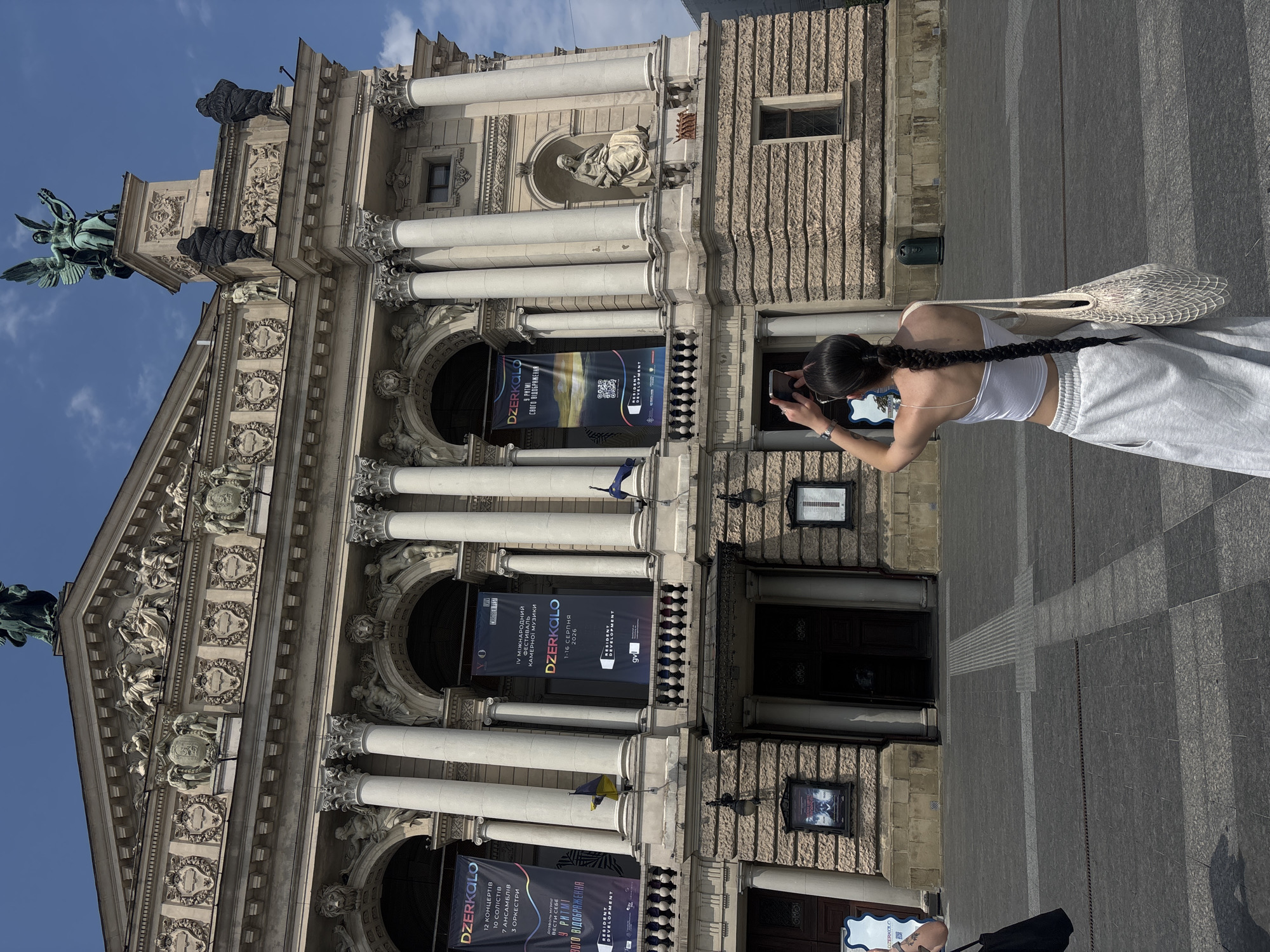 This is a medium shot of a woman with a long braid, wearing a white tank top and grey sweatpants, taking a picture of a building with her phone. She is standing in front of the Lviv Opera House, a grand Neorenaissance building in Lviv, Ukraine. The building is adorned with statues, columns, and ornate carvings. Two large banners hang from the facade of the building, advertising events. A Ukrainian flag is visible on the left side of the building. The ground in front of the building is paved with dark grey tiles arranged in a grid pattern. The sky is blue with some scattered clouds. The overall scene is a bright, sunny day in a public square.