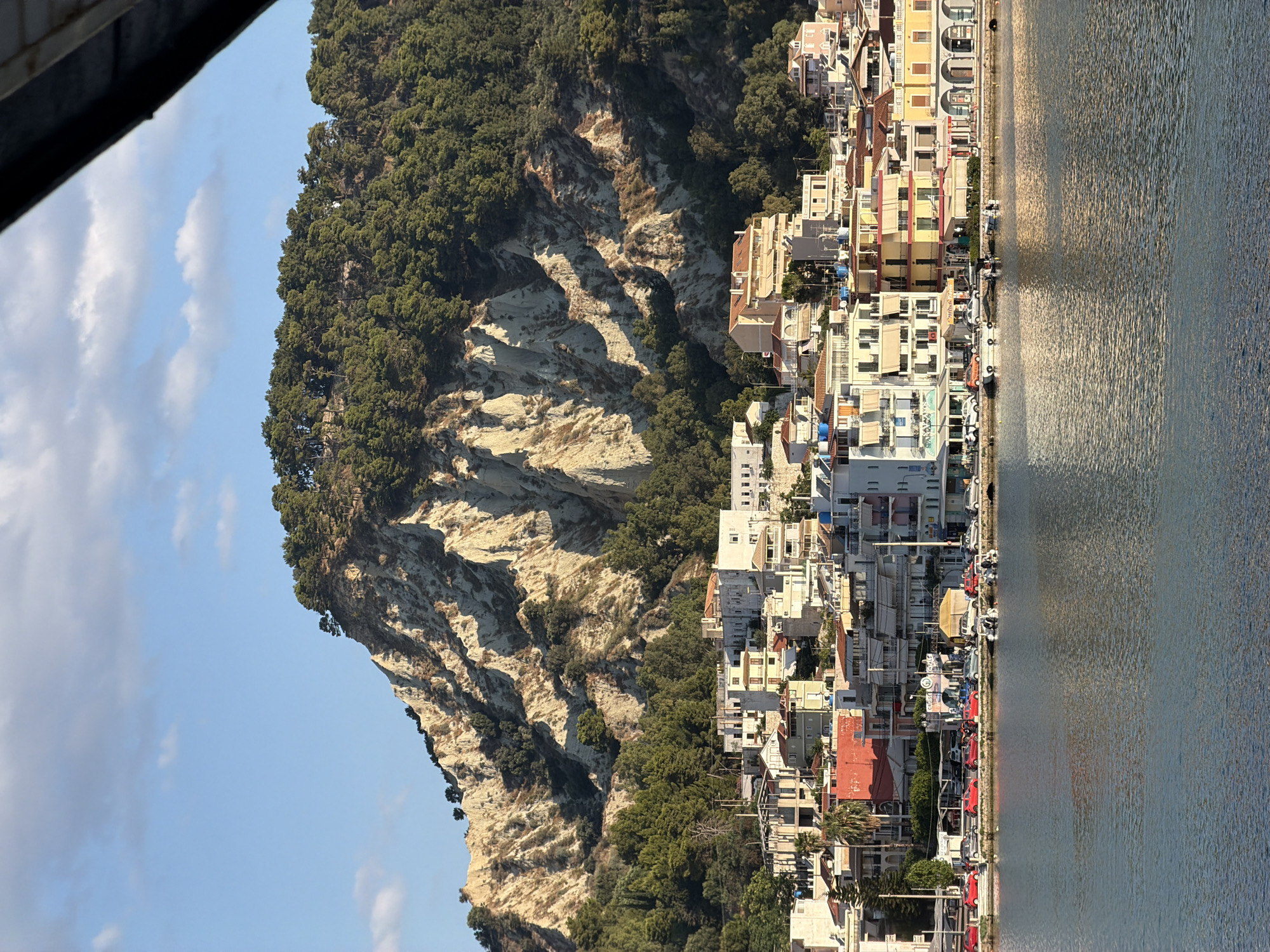 The image shows a coastal town with buildings lining a waterfront promenade. Behind the town, a steep, rocky hill is covered with dense green trees. The water in the foreground is calm and reflects the sunlight. The buildings are colorful, with a mix of white, yellow, and red facades, and many have balconies. Cars are parked along the street bordering the waterfront. A few small boats are docked in the water. The sky is blue with some scattered white clouds. The scene suggests a sunny day in a Mediterranean coastal town, likely Zakynthos town on the island of Zakynthos, Greece, given the characteristic landscape and architecture.