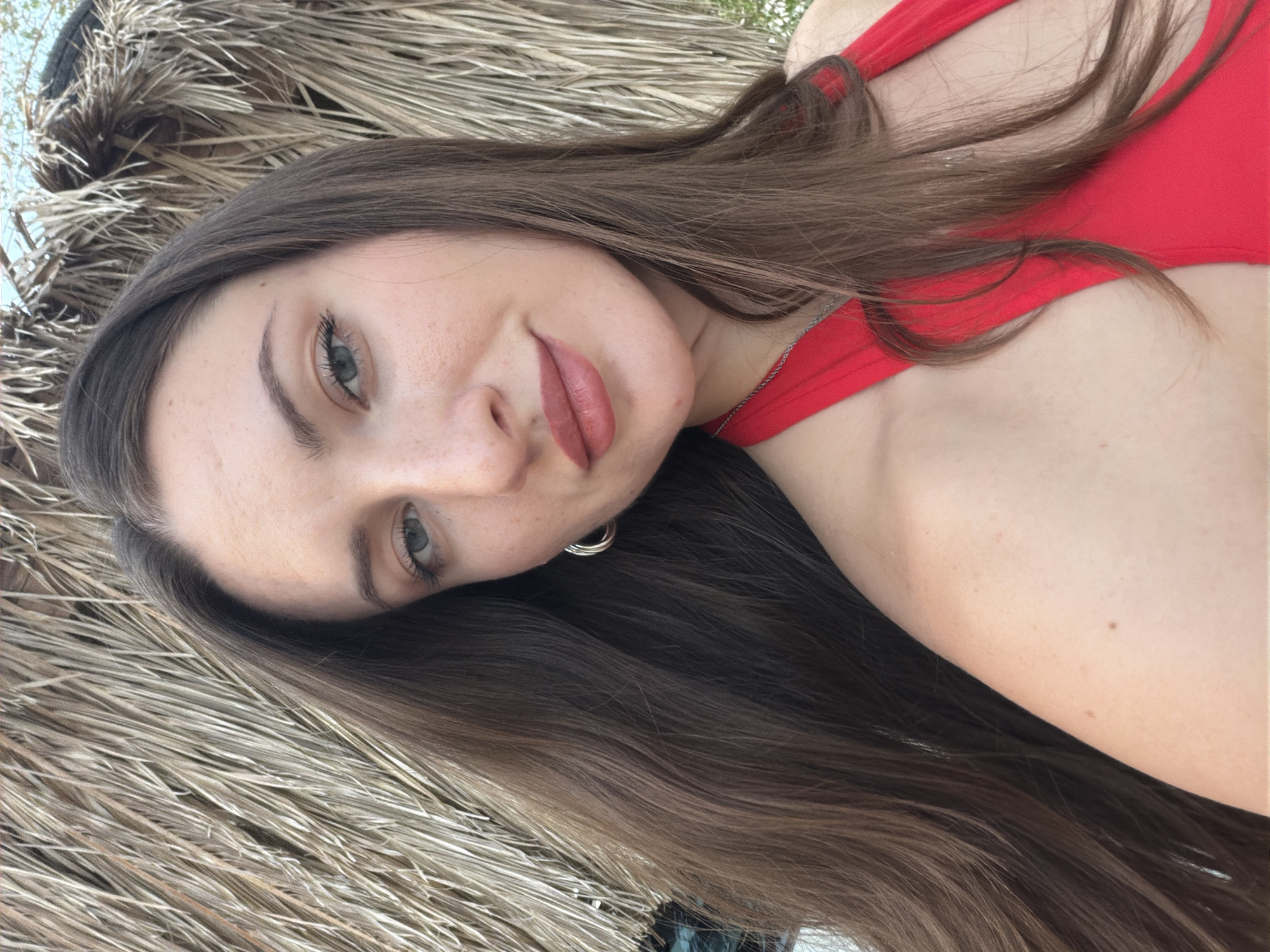 A young woman with long brown hair, wearing a red halter top and silver hoop earrings, poses for a selfie. Her eyes are light blue, and she is wearing subtle makeup. She is positioned in front of a thatched roof structure, common in beach resorts in Sharm El Sheikh, Egypt, suggesting an outdoor setting possibly near a beach or pool. The lighting indicates it is daytime.