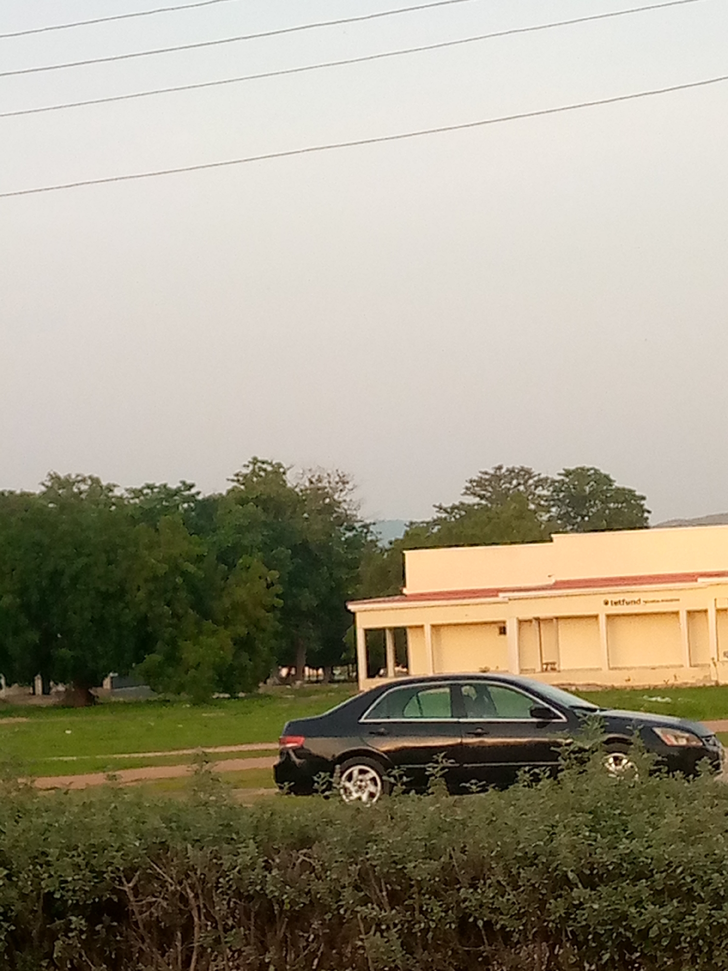 A black sedan is parked on a dirt road in front of a light-colored building with arched openings. Lush green trees and bushes are visible in the foreground and background. A sign with "tetfund" is affixed to the building. The sky is overcast.