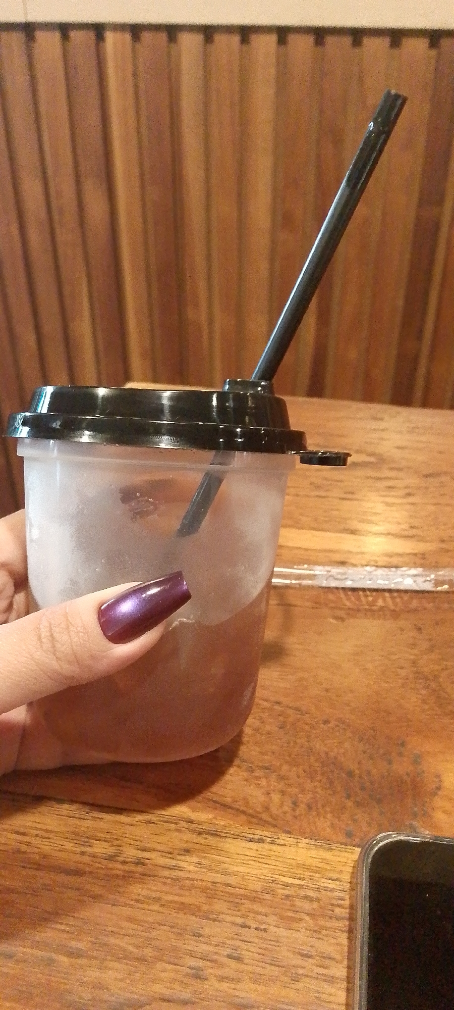 A medium shot shows a person's hand, with a dark purple, glossy nail polish, holding a clear plastic cup filled with iced tea. The cup has a black lid and a black straw. The background is a wooden wall with vertical planks and a wooden table. A smartphone is partially visible in the lower right corner of the image. The scene appears to be indoors, possibly at a cafe or restaurant.