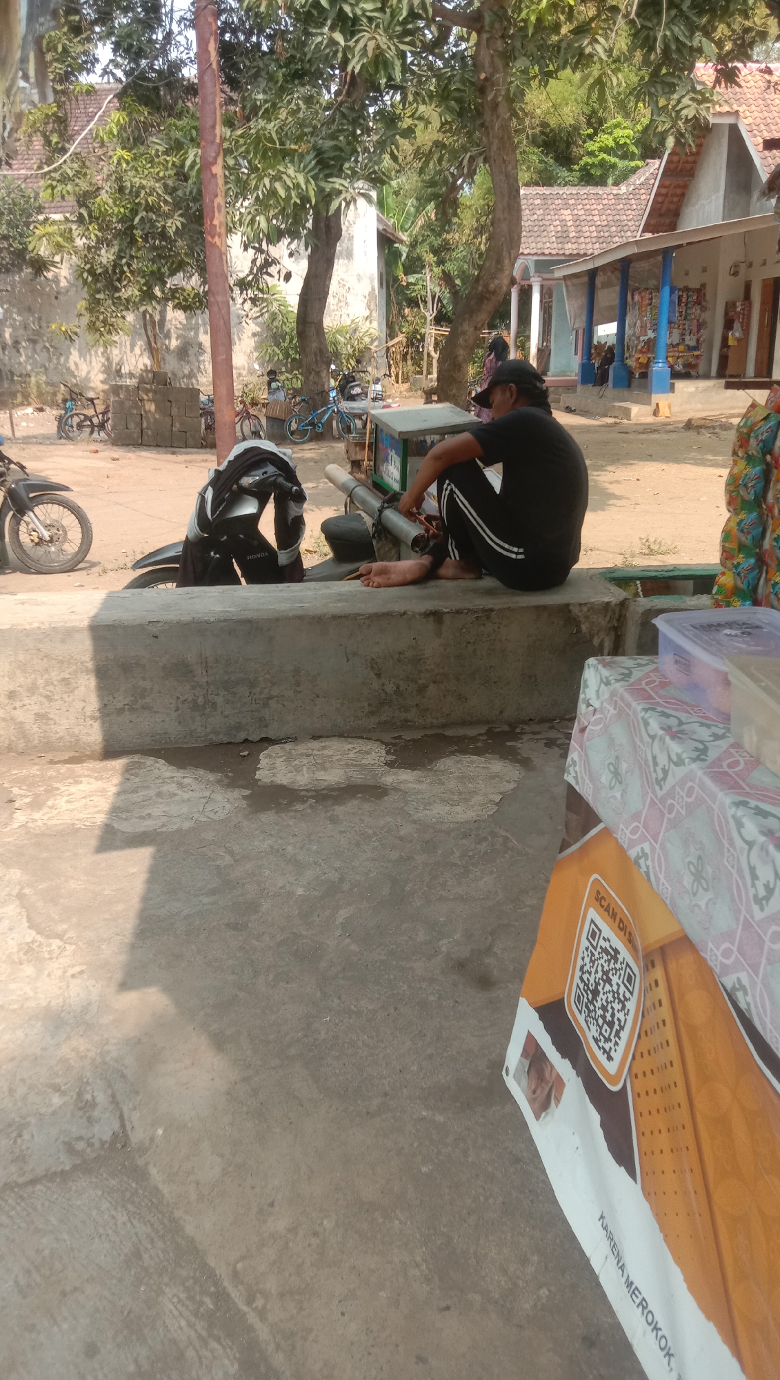 In Ngempit, Indonesia, a person wearing a black t-shirt and black pants with white stripes is sitting on a concrete ledge. They are facing away from the camera, looking towards a street scene. A motorcycle with a black and silver helmet on its handlebars is parked behind the person. In the background, a small building with a tiled roof and blue pillars is visible, possibly a shop. Several bicycles are also parked near the building. The ground is paved with concrete and dirt. The overall atmosphere is casual, with the person appearing to be resting or observing their surroundings.