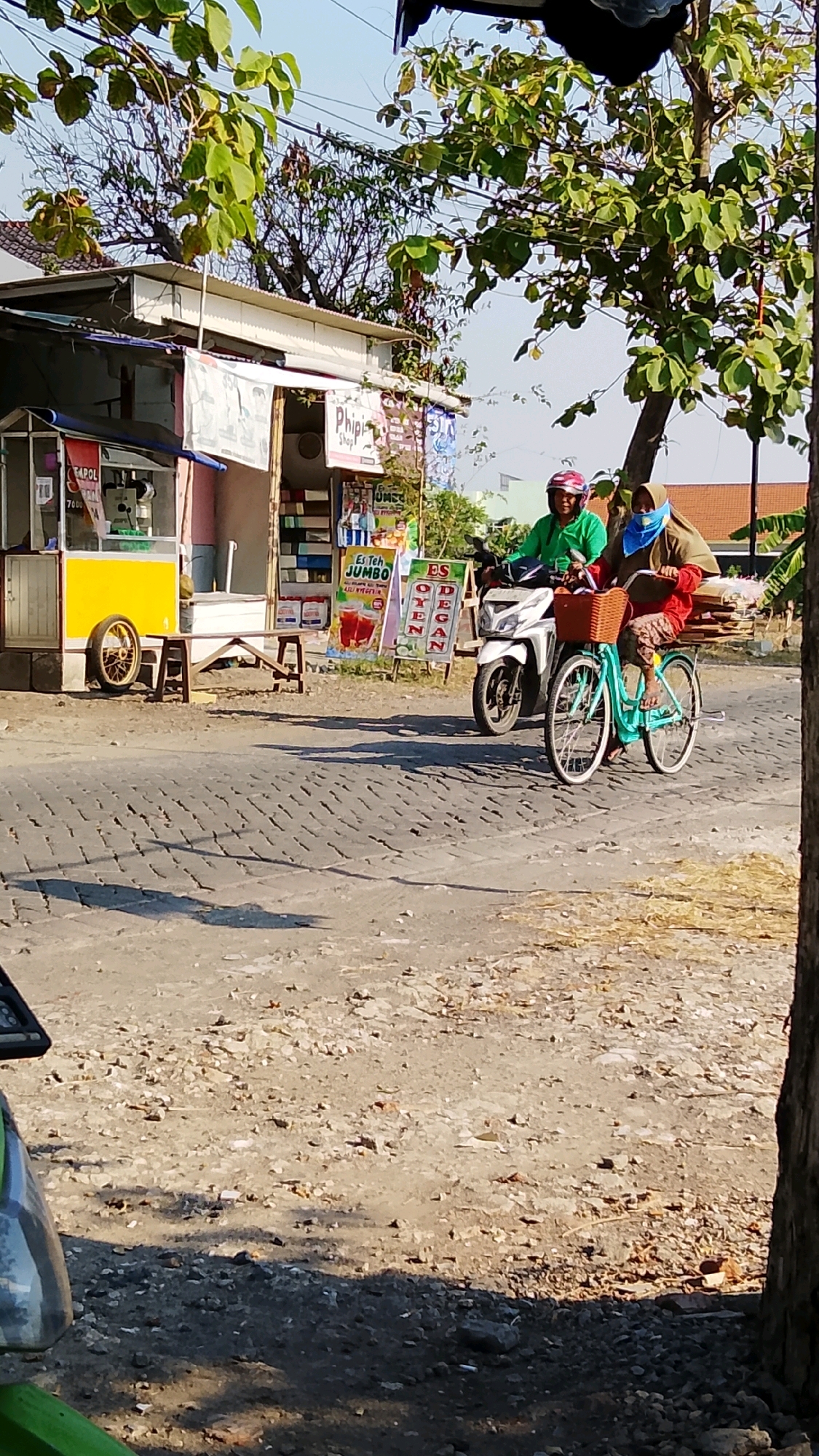 In Dahanrejo, Indonesia, two individuals are captured in a daytime scene on a street. A person wearing a green long-sleeved shirt and a red head covering is riding a white motorcycle, with another person in a red shirt and a light brown head covering seated behind them. The passenger is also wearing a blue face covering and has a basket with items on the front of a turquoise bicycle parked in front of the motorcycle. The street appears to be paved with cobblestones, and there are shadows cast by trees and overhead wires. To the left of the scene, there is a small roadside stall with a yellow base, a white roof, and signs indicating it sells beverages. One sign displays an image of iced tea and reads "Es Teh Jumbo." Another sign features the word "ES" vertically. The environment suggests a commercial area with food and drink vendors.