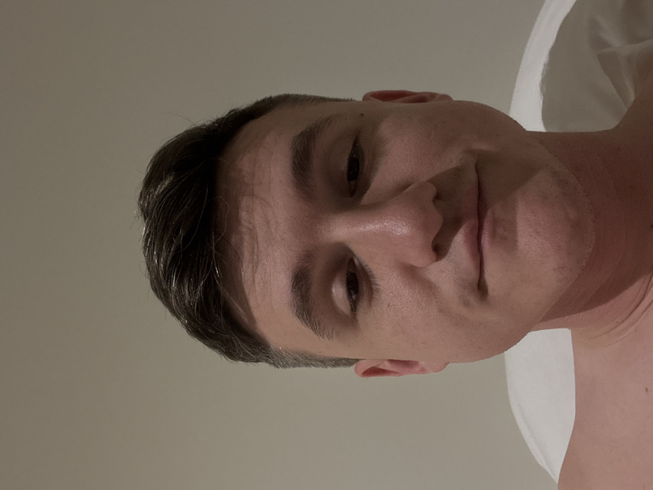 The image presents a close-up, frontal portrait of an adult male. He possesses short, dark brown hair, light skin, and a light stubble on his jawline and upper lip. His eyes are directed downwards and partially closed, while his lips form a slight, closed-mouth smirk. The subject's bare left shoulder is visible in the lower left of the frame. A white pillow is partially visible behind his right shoulder, suggesting an indoor environment, likely a bed. The background is uniformly light-colored and featureless, providing no identifiable architectural or environmental details. The specific location within the city of Venice, Italy, cannot be determined from the visual evidence presented in the image.
