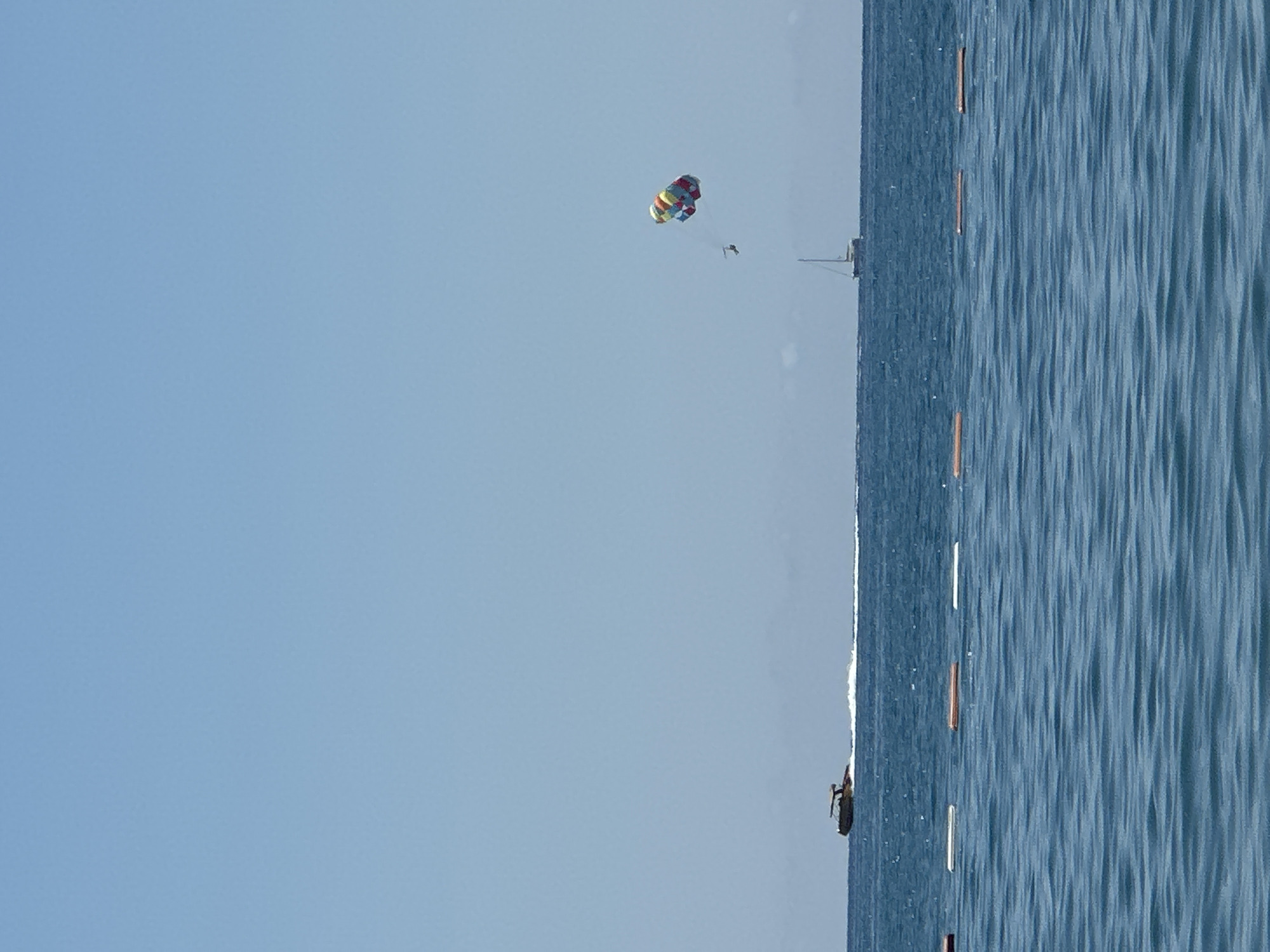 A wide-angle, eye-level, outdoor shot depicts a bright, clear sky over a tranquil blue sea, with a distinct line of buoys in the foreground. In the middle ground, a speed boat creates a white wake as it travels across the water. Further out, a sailboat is visible on the horizon, and above it, a person is parasailing, their colorful parachute a vibrant splash against the blue sky. The scene appears to be set in the vicinity of Boreti, Montenegro, a location known for its coastal activities and clear waters.