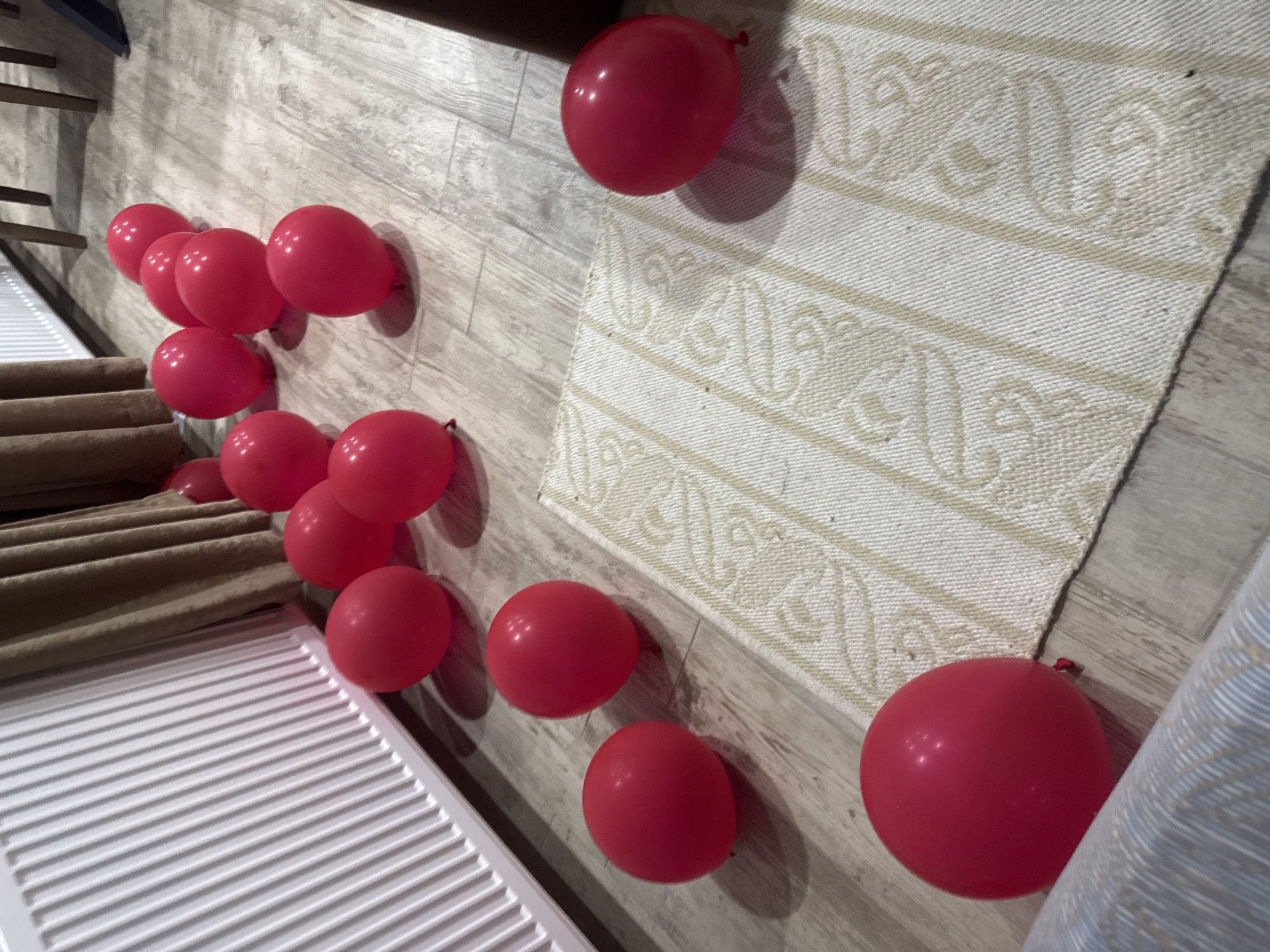 The scene shows a section of a room, possibly in Lehchevo, Bulgaria, although there is no confirmation of this location within the image itself. The floor has a light wood-grain pattern, upon which red balloons are scattered, seemingly leading away from a staircase with a white railing and tan carpeting. Adjacent to the balloons is a patterned rug and the edge of a patterned drape or sheet. No people are visible. The scene suggests a celebratory event or decoration, without providing further context.