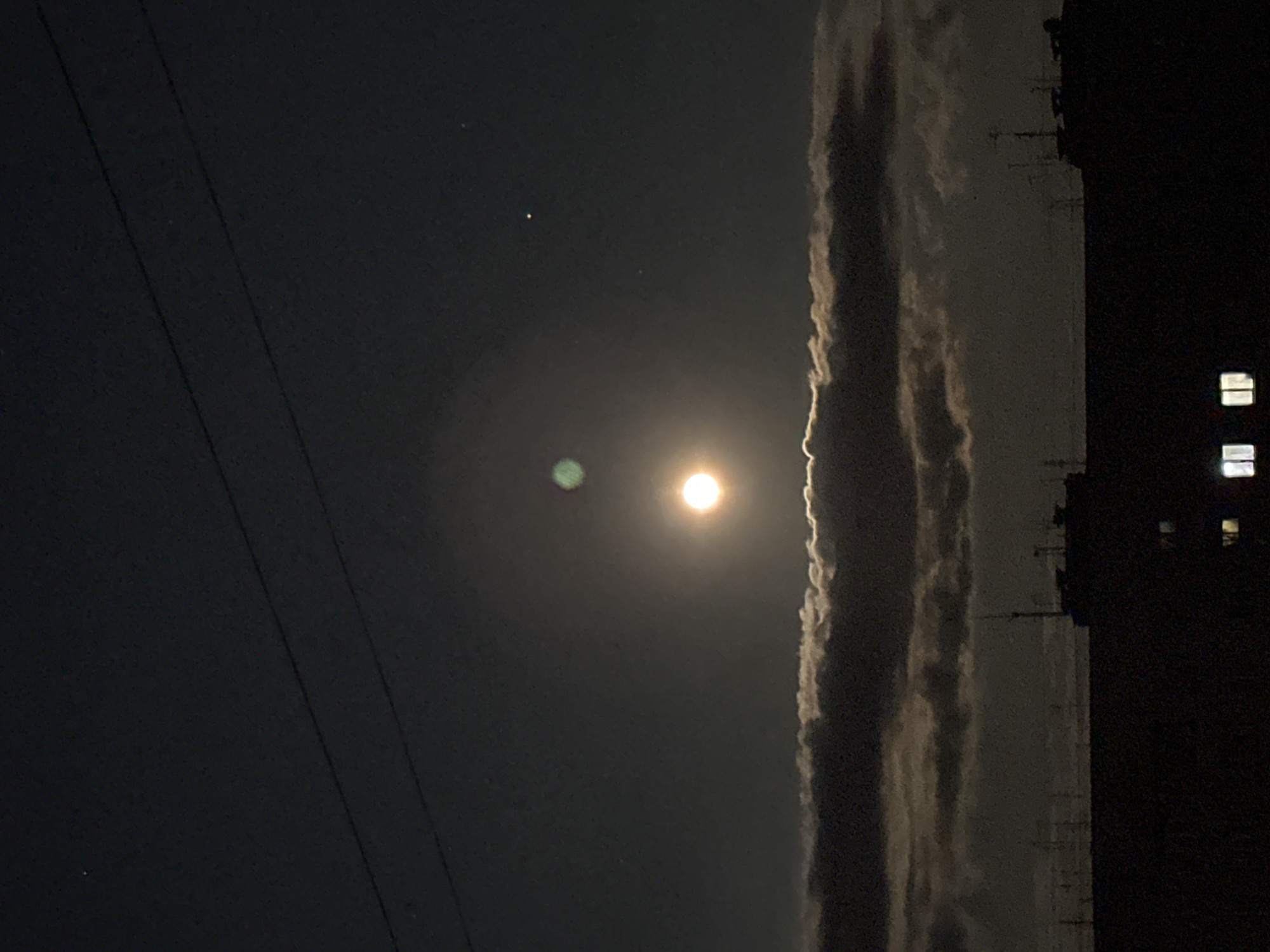 The image depicts a night scene, likely in an urban environment. A bright light source, probably the moon, dominates the center, with a circular halo effect around it. To the right of the light source, a dense formation of clouds is visible, illuminated by the bright light. Further to the right, a dark structure, possibly a building, is silhouetted against the sky, with a few lit windows. Dark wires are seen on the left side of the image. The overall environment appears to be dimly lit, and the image is rotated 90 degrees counter-clockwise.