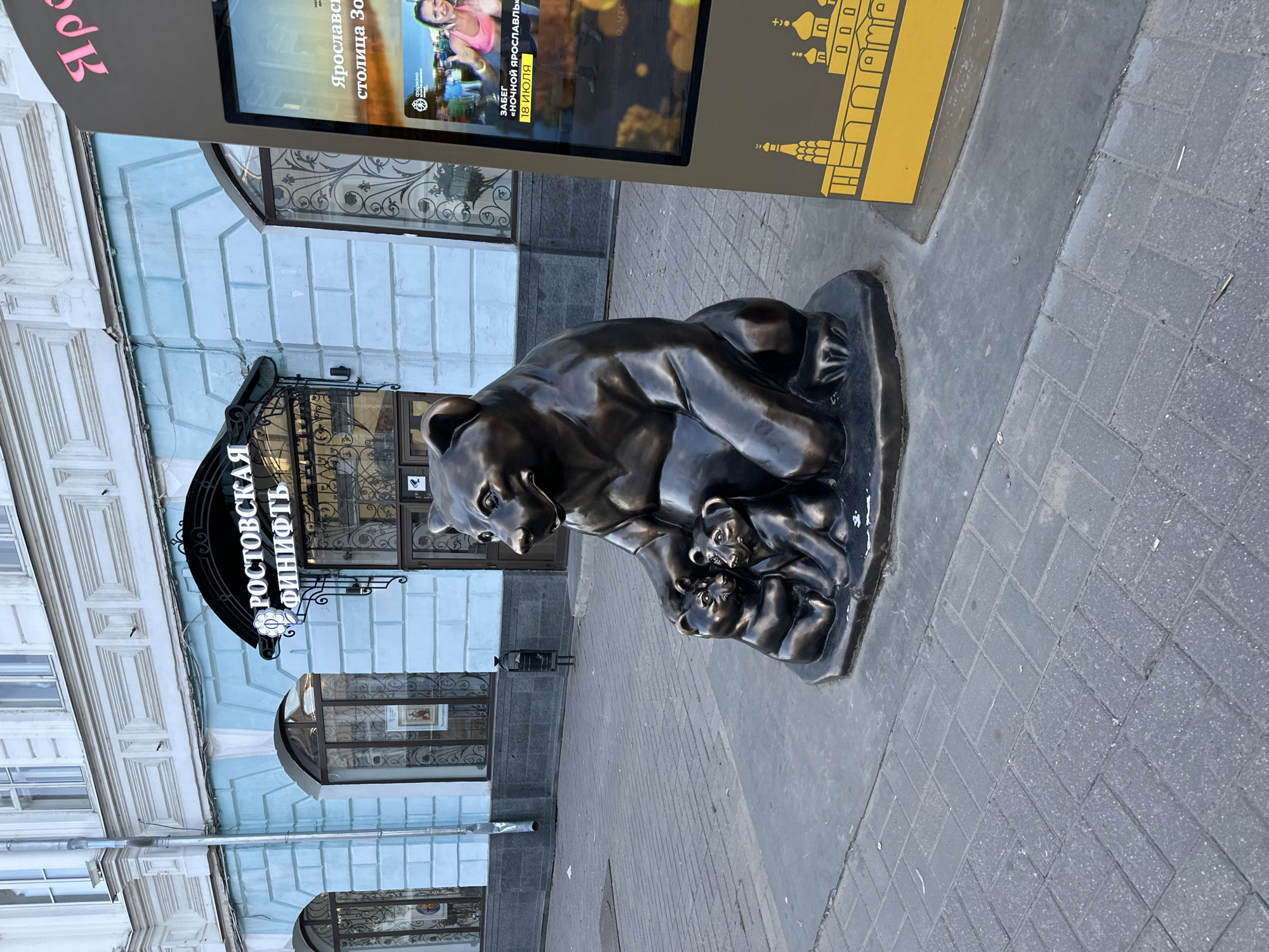 The image shows a bronze sculpture of a mother bear with two cubs, located in front of a building with light blue and white painted facade. Above the entrance of the building, there is a black awning with white Cyrillic text that reads "РОСТОВСКАЯ ФИНИФТЬ". To the right of the sculpture and building, a digital billboard displays an advertisement. The billboard shows a woman participating in a race and text in Russian. The ground in front of the sculpture and building is paved with grey cobblestones. The scene appears to be a street or pedestrian area in a city.