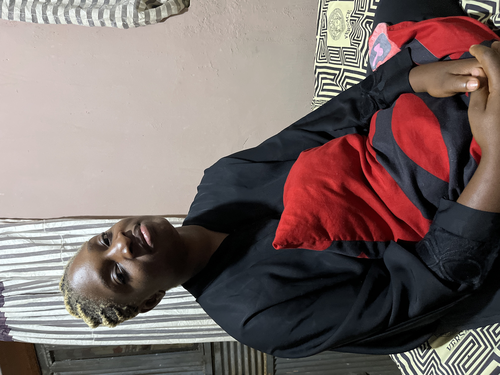 A young Black woman with blonde-streaked braided hair is seated, holding a red and black patterned pillow. She is wearing a loose-fitting black garment. The background consists of a light-colored wall, a window with striped curtains, and a patterned bedspread. The scene appears to be indoors, possibly in a residential setting in Rafin Par, Nigeria.
