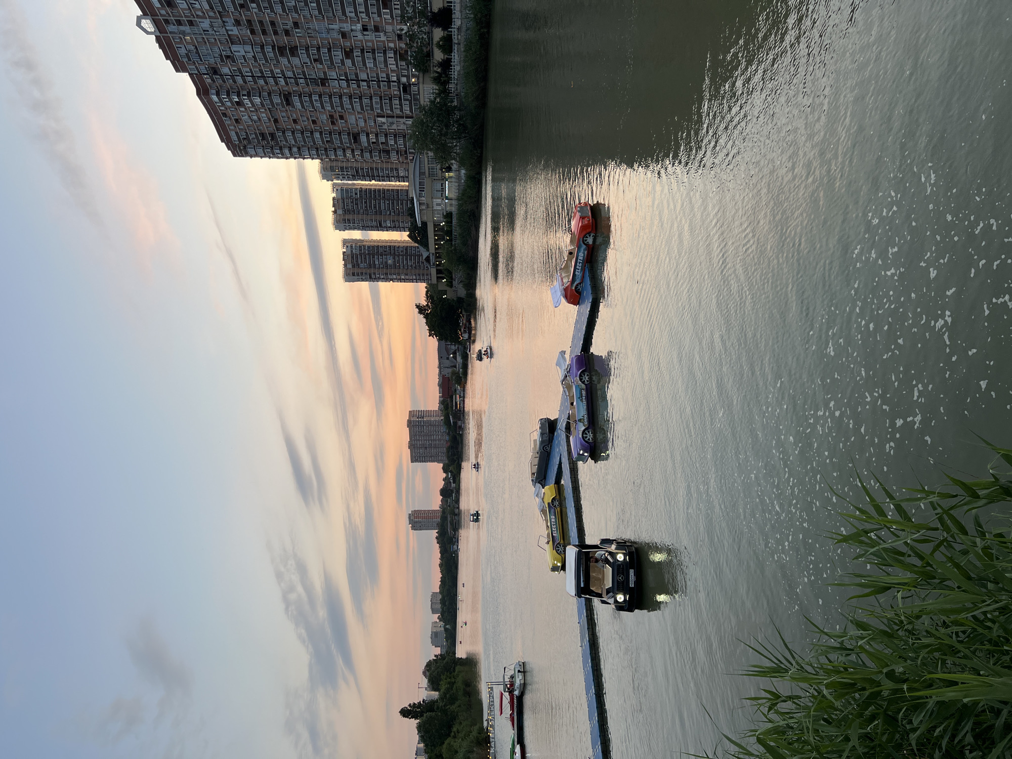 At sunset, on the Krasnodar river in Krasnodar, Russia, the sky displays a gradient of soft pinks, oranges, and blues, with scattered wispy clouds. The river is calm, reflecting the pastel hues of the sky and the city lights. Several uniquely shaped pedal boats, designed to resemble vintage cars, are docked or floating on the water. One black car-shaped pedal boat, with its headlights on, is being operated by a person. Further out on the river, other small boats and pedal boats can be seen. Along the riverbanks, lush green vegetation is visible in the foreground on the left, and trees line the opposite bank. In the background, modern high-rise apartment buildings dominate the cityscape, their windows reflecting the evening light. The overall scene suggests a peaceful recreational area in an urban setting.