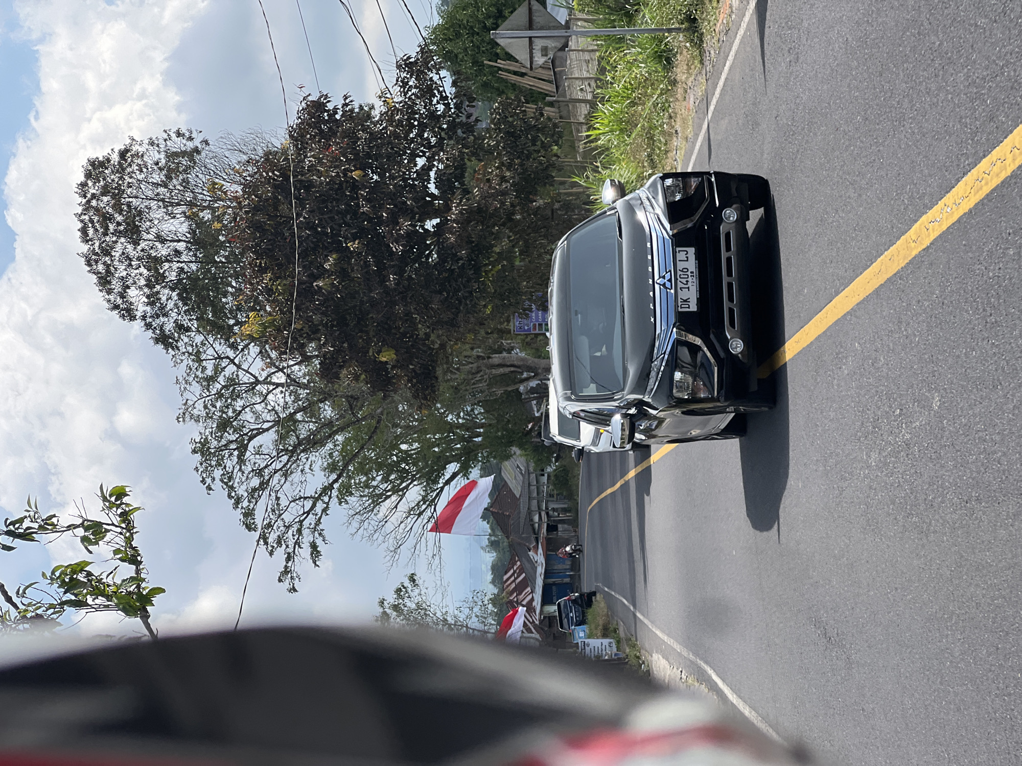 The image depicts a view from a vehicle, likely a motorcycle given the partial view of a helmet in the foreground, traveling on a paved road in Kintamani, Indonesia. The road has a distinctive yellow line marking its center. In the mid-ground, several vehicles are visible, including a black Mitsubishi Xpander with license plate DK 1406 LJ prominently displayed, and a white SUV. Further ahead, a person is seen riding a motorcycle. On the left side of the road, there are several Indonesian flags (red and white) hoisted, indicating a potential national celebration or event. Buildings with corrugated metal roofs line the left side of the road, suggesting a rural or semi-urban setting. Lush green vegetation and trees, some with dark foliage, surround the road. The sky is partly cloudy with a bright, daytime light. The background reveals a hilly or mountainous landscape, characteristic of Kintamani.
